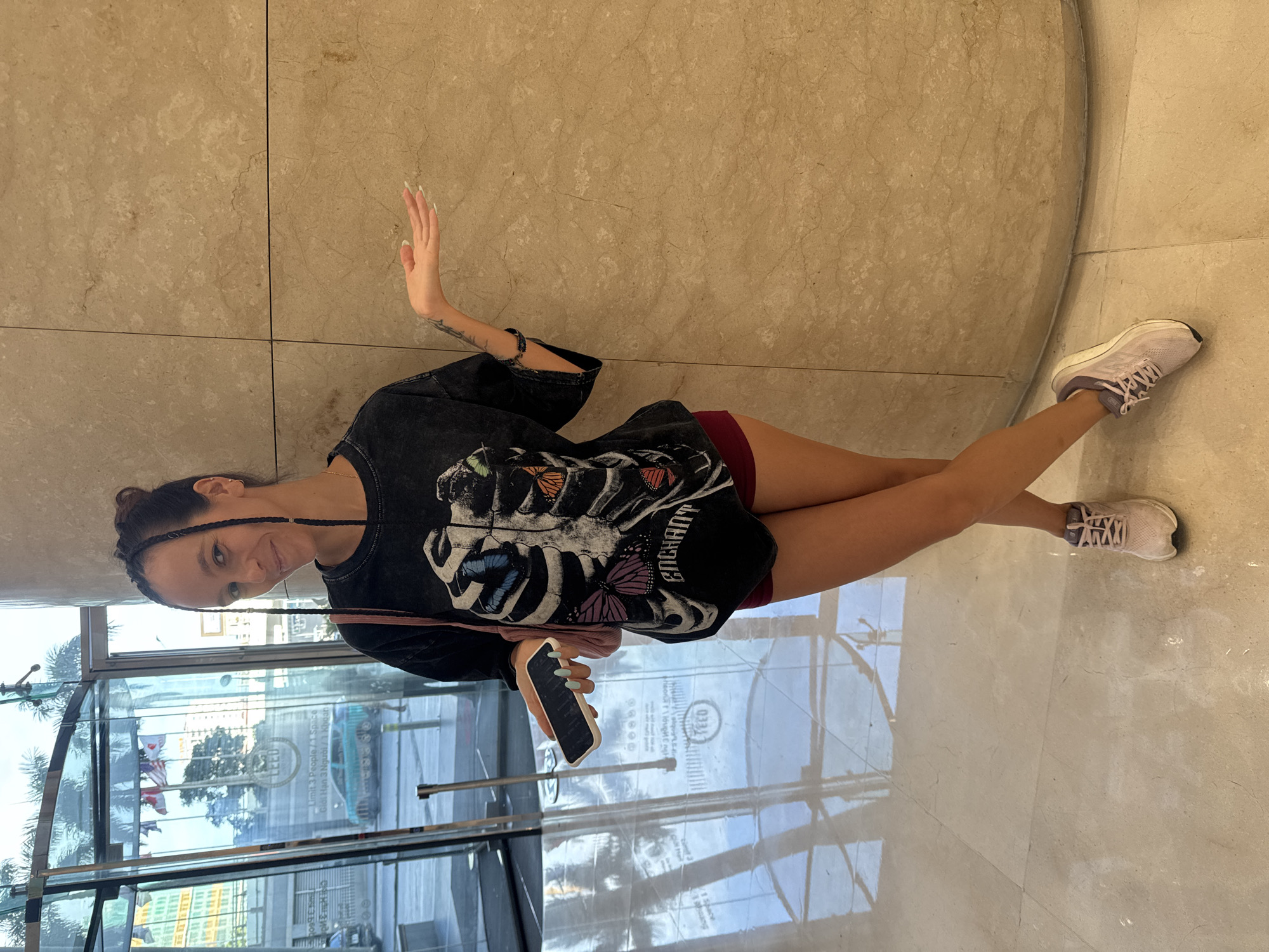 A woman stands in a tiled lobby with a marble column on the right. She has dark braided hair pulled up and is wearing a black t-shirt with a skeleton ribcage and butterfly graphic, burgundy shorts, and light pink sneakers. She holds a white phone in her left hand and her right hand is extended outwards. In the background, through a glass revolving door, trees, flags, and buildings are visible. The floor has a reflective surface.