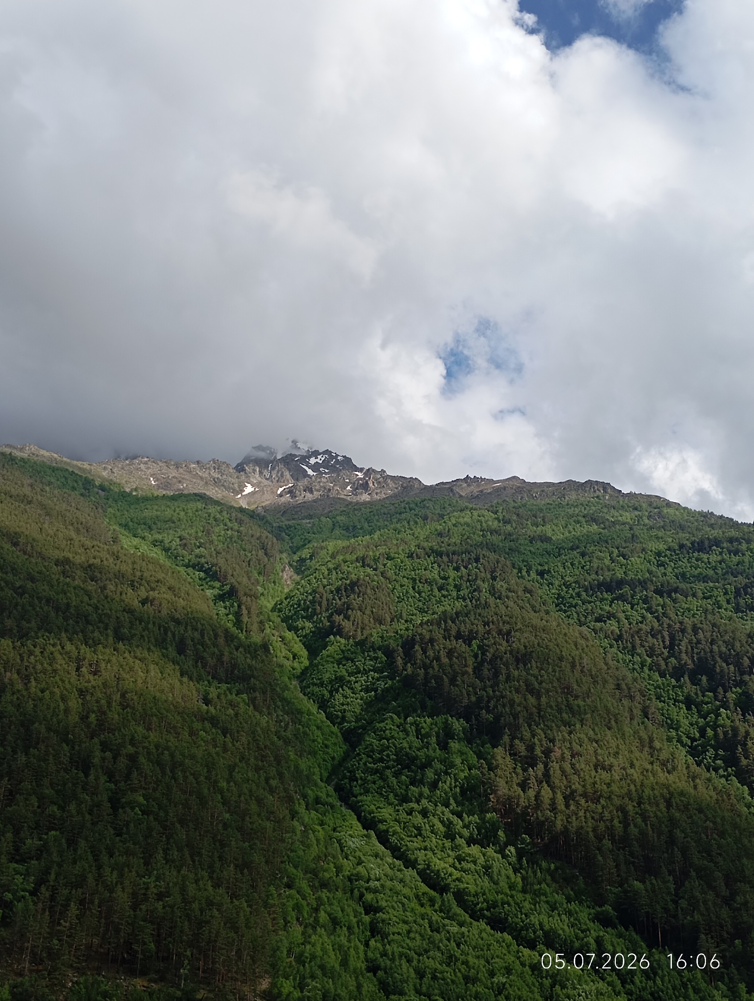 The image shows a mountainous landscape with a dense forest covering the slopes. The peaks of the mountains are rocky and partially covered in snow. The sky is cloudy with some patches of blue. A timestamp "05.07.2026 16:06" is visible in the lower right corner of the image. The location is identified as the vicinity of the city of Neutrino, Russia. The scene is devoid of any people or man-made structures, focusing solely on the natural environment.