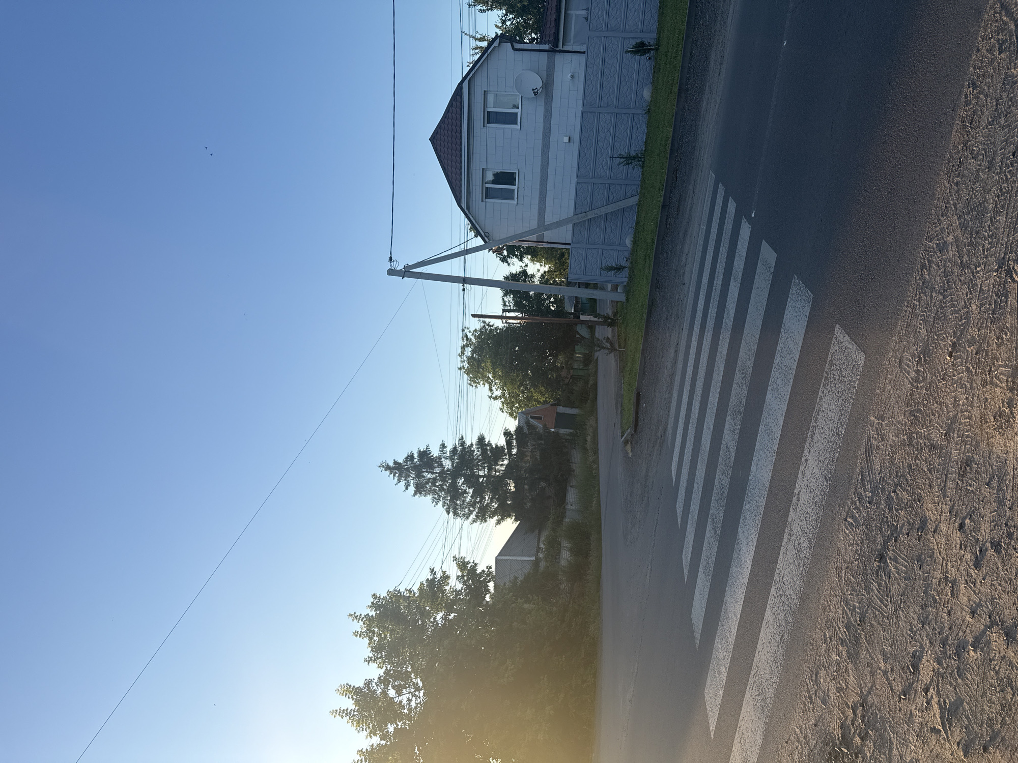 The image shows a pedestrian crossing on an asphalt road in a suburban area. The road is bordered by a dirt shoulder on the left and grass and a fence on the right. Behind the fence, a two-story house with a gray tiled roof and white walls is visible, with a satellite dish mounted on the side. Several utility poles and wires run across the scene, and various trees are present, including a tall evergreen. The sky is clear and blue, indicating daytime. The presence of a pedestrian crossing suggests a location within a populated area, likely a residential street or an intersection. Considering the context provided, this scene is located in the city of Dnipro, Ukraine. There are no people visible in the image.