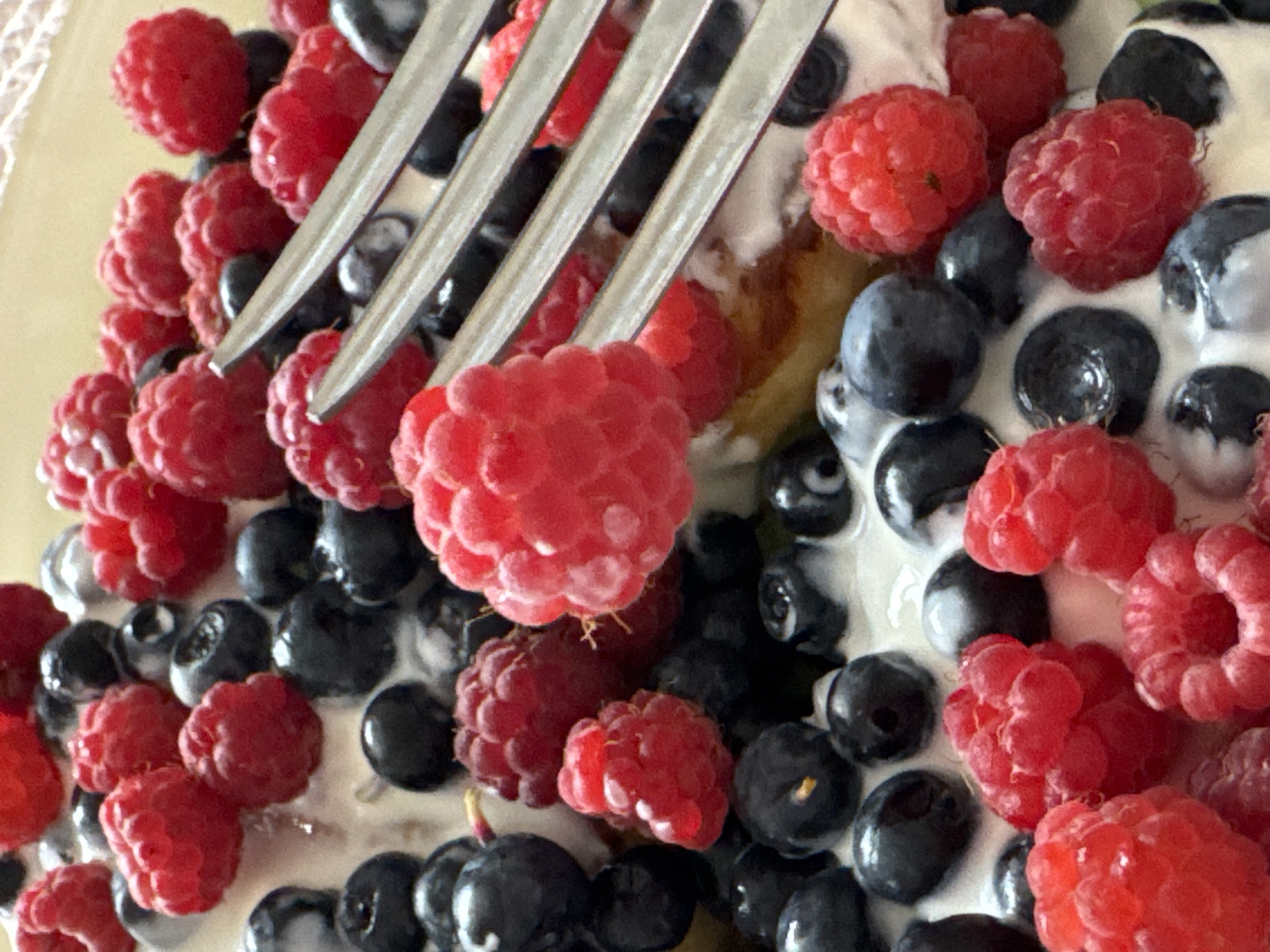 A close-up shot captures a serving of fresh berries, including raspberries and blueberries, atop a creamy white base, possibly yogurt or whipped cream. The berries are arranged in a dense cluster, with the vibrant red raspberries and deep blue blueberries creating a visually appealing contrast. The texture of the raspberries, with their individual drupelets, is clearly visible, as are the smooth, round surfaces of the blueberries. A portion of a fork with three tines is present in the upper right of the frame, appearing to rest on top of the berries, suggesting the food is ready to be eaten. The background is a soft, light yellow, creating a simple and clean backdrop that emphasizes the colorful fruit. The image is taken from a high angle, looking directly down onto the dish. There is no indication of the city of Ladozhskoe Ozero, Russia, or any specific environmental details beyond the food itself.
