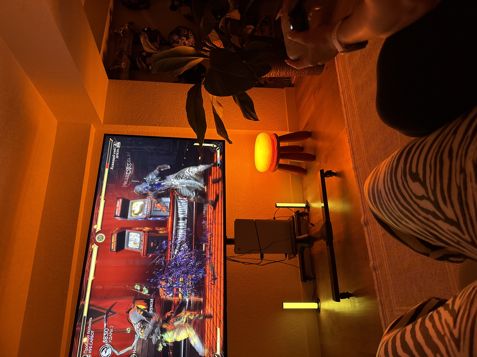 The image shows a dimly lit room with a large television displaying a fighting video game, likely Mortal Kombat. The room is illuminated by warm, orange ambient lighting, creating a cozy atmosphere. A plant with large leaves sits to the right of the television, and a small red stool with a glowing yellow top is placed in front of it. In the foreground, a person's legs are visible, wearing patterned pants, suggesting they are sitting or lounging. Another person's hands are also visible, holding a video game controller, indicating active gameplay. A game console is positioned below the television. The overall scene suggests a relaxed evening spent gaming in a residential setting. The location of this scene in Yerevan, Armenia, cannot be determined from the provided image.