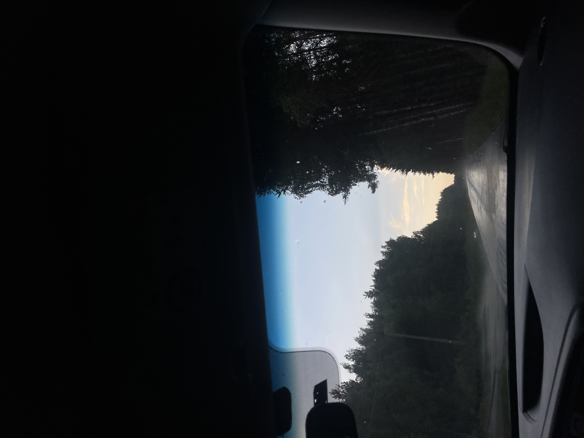 The image, taken from inside a vehicle, shows a view through the windshield of a wet asphalt road through a forest. The sky is overcast with a hint of sunset colors on the horizon. The road appears to be in a rural area, with trees lining both sides. There are no people or other vehicles visible in the scene. The dashboard of the car is partially visible in the foreground. The location is identified as Pokrovskoye, Russia.