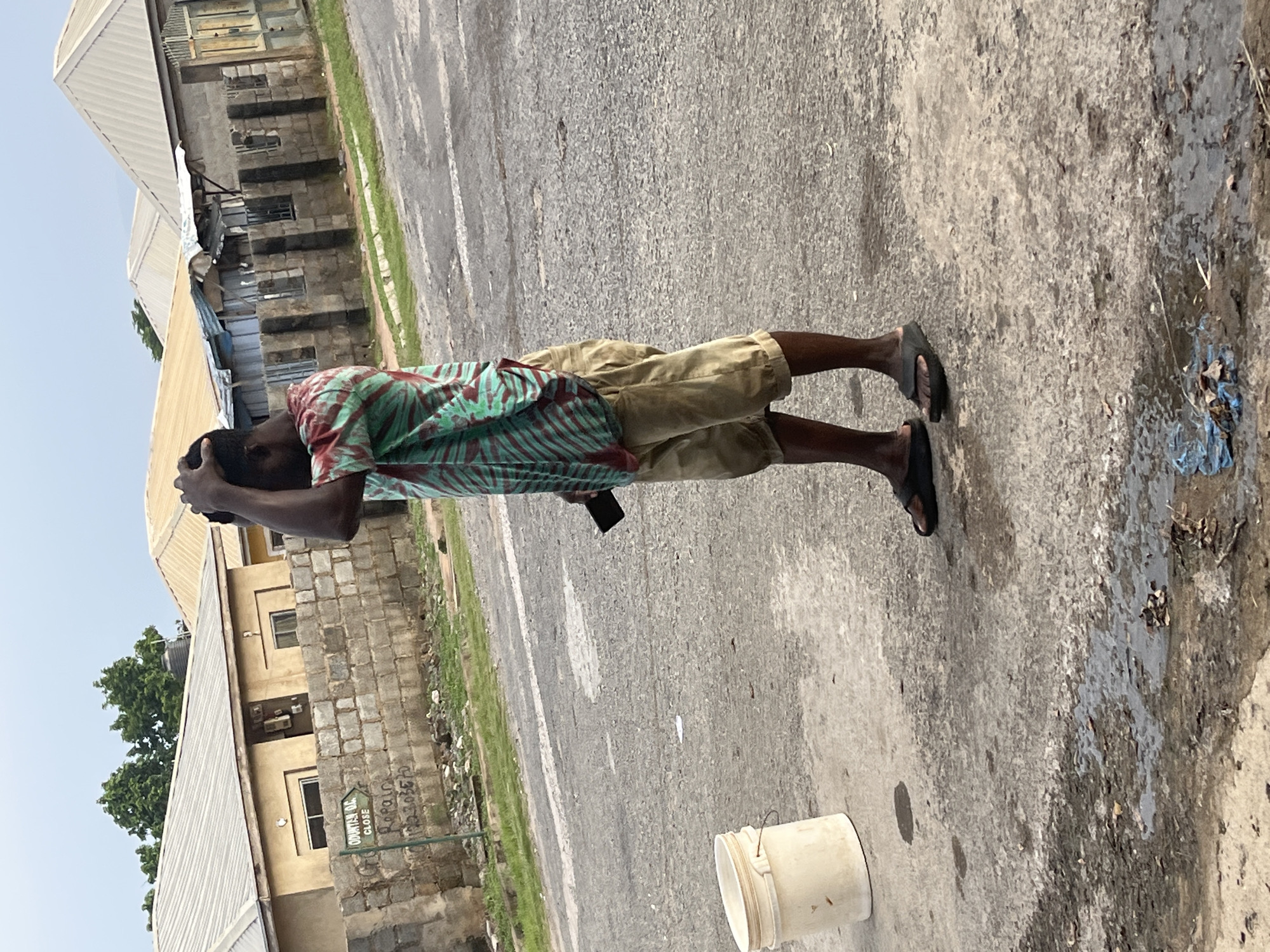 In Abuja, Nigeria, a person stands on a paved road, facing left. They are wearing a red and green striped short-sleeved shirt and tan shorts. The person has their right hand on their head. In their left hand, they hold a dark rectangular object, possibly a phone. A white plastic bucket is to the left of the person. In the background, several buildings with corrugated metal roofs are visible, along with a brick wall and a street sign. The environment appears to be a residential area in the city.