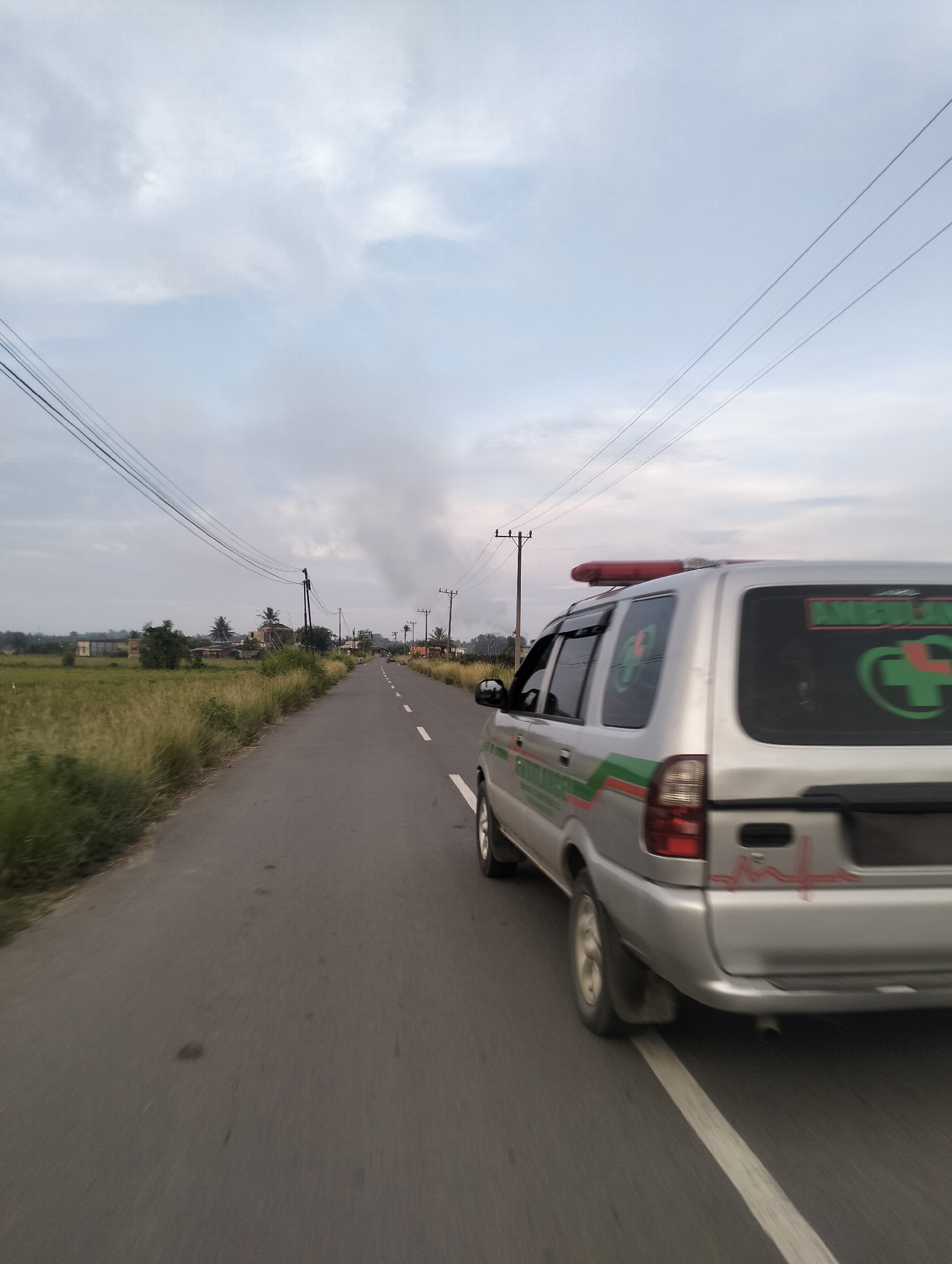 In Muliengmanyang, Indonesia, an ambulance vehicle, a silver SUV with green and red markings, travels down a paved road lined with grassy fields and scattered buildings. The ambulance features a red emergency light bar on its roof and has a green cross emblem on its rear window. Power lines crisscross the sky, and utility poles are visible along the roadside. The sky is overcast with clouds, indicating a potentially gloomy day. The road is marked with a white dashed line, and the perspective suggests the image was taken from another moving vehicle.