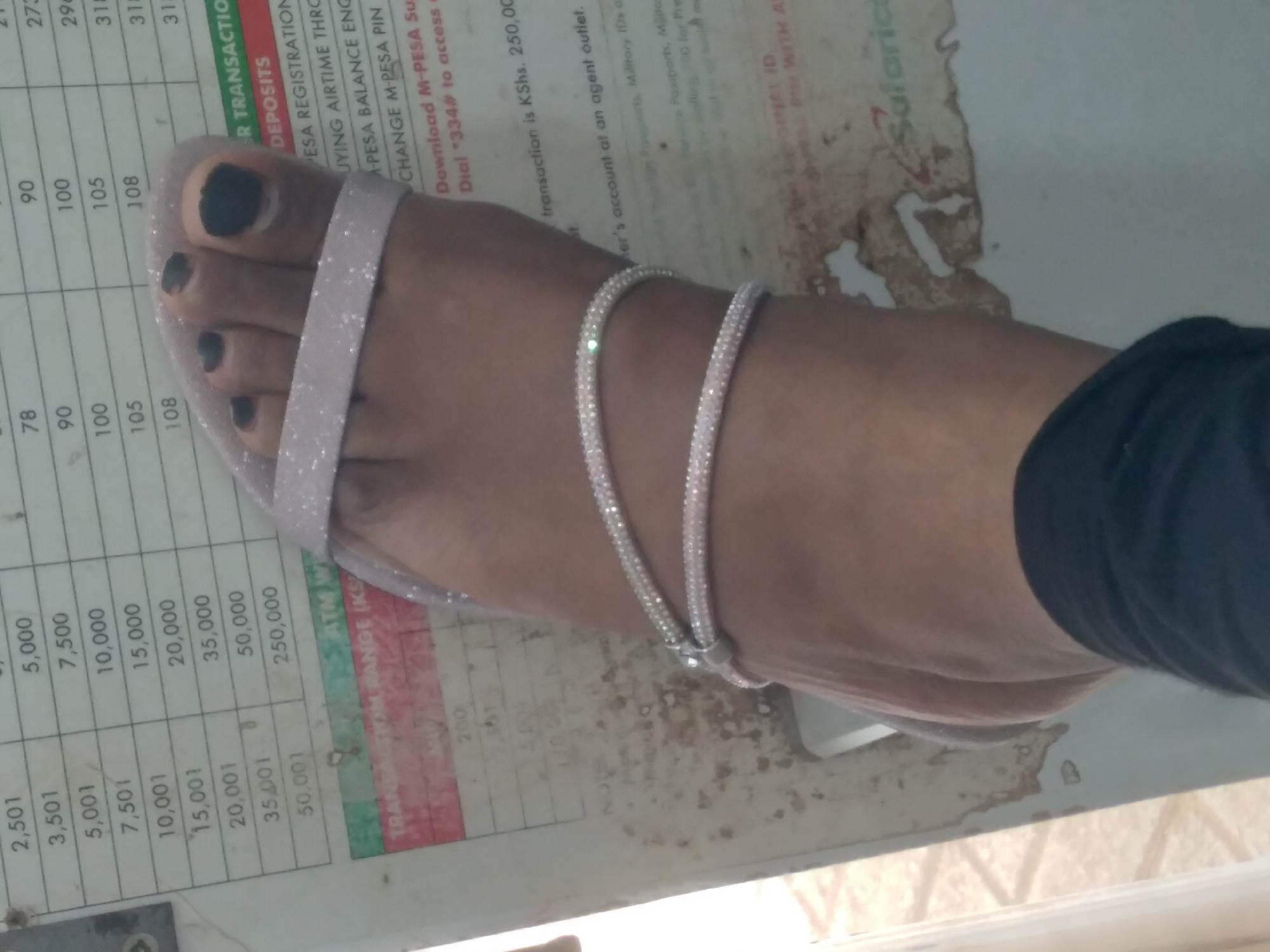 A person's left foot with black nail polish is shown. The foot is wearing two sparkling silver anklets. The background consists of a table with numbers and text, possibly a price list or fee schedule. Part of the text visible includes "TRANSACTION RANGE (KSh", "DEPOSITS", and "SAFARICOM". The overall scene suggests an indoor environment with the foot resting on a surface. The presence of Safaricom branding indicates the location is likely in Kenya, and given the provided context of Nyeri, Kenya, it is plausible the image was captured there.