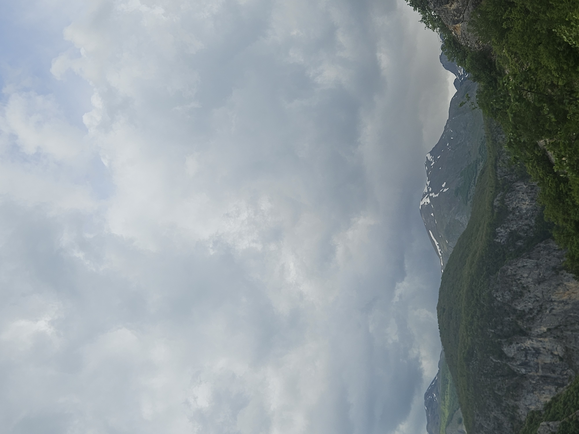 The image depicts a mountain landscape in Plužine, Montenegro, under an overcast sky filled with grey and white clouds. The mountains are rugged, with visible rock formations and green vegetation covering their slopes. Some areas exhibit patches of snow, indicating higher elevations. The perspective is slightly angled, suggesting the photo may have been taken from a lower vantage point looking upwards.