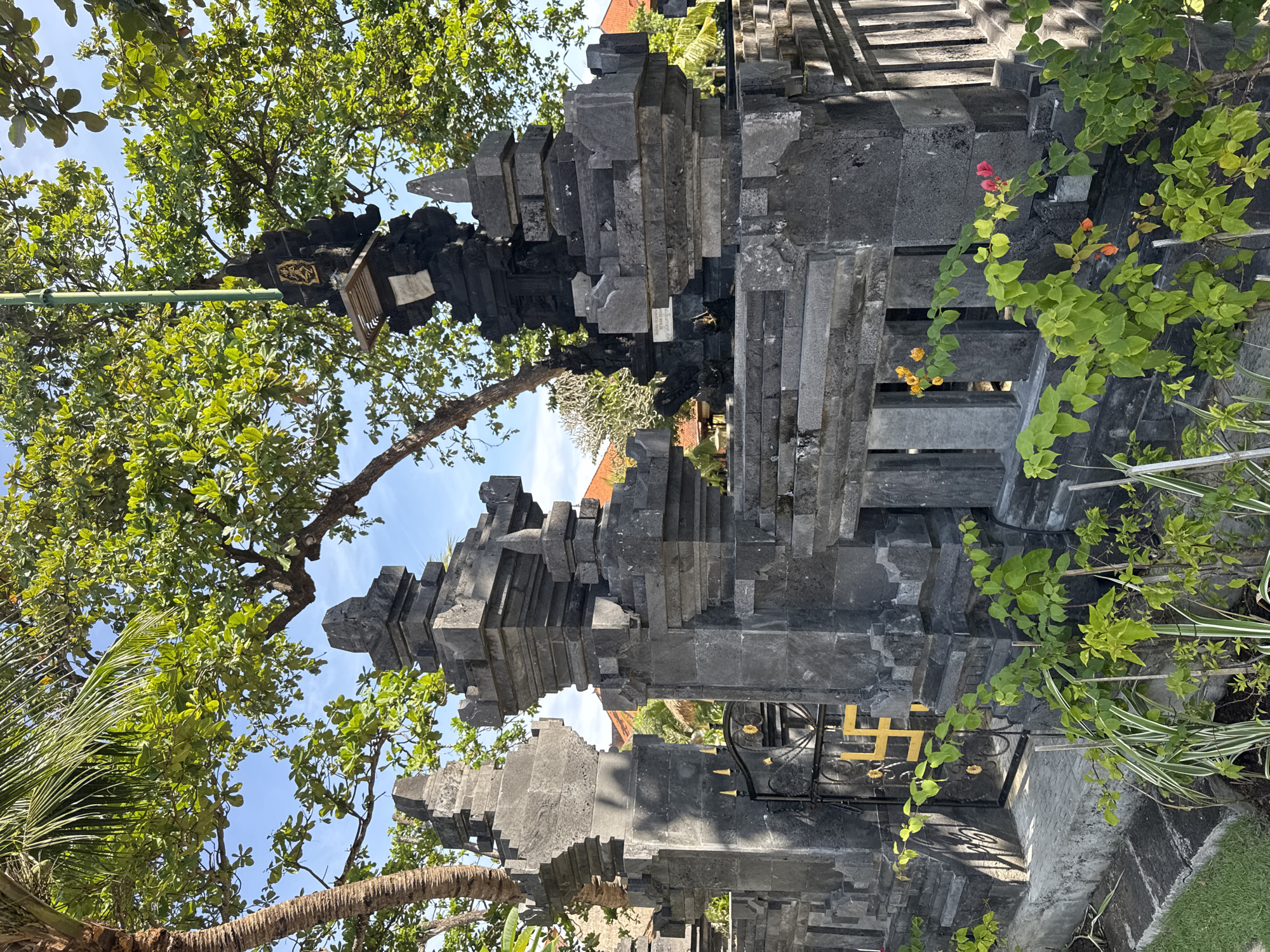 The image depicts a stone structure, likely part of a traditional Balinese temple or gate, in Kuta Selatan, Indonesia. The structure is made of dark gray stone blocks arranged in a tiered, stepped design. It features a black iron gate with golden details. Lush green vegetation, including trees with broad leaves, flowering vines with red and orange blossoms, and ornamental grasses, surrounds the structure. The sky is visible through the trees and structures, appearing blue with some clouds.