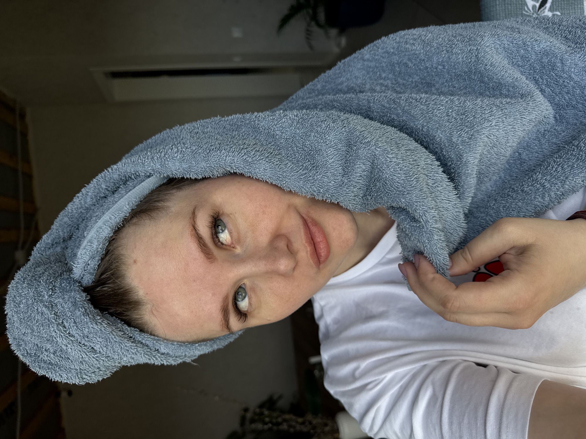 The image is oriented on its side, depicting a young woman seen from the chest up, looking directly at the viewer. She has fair skin, prominent blue-grey eyes, dark eyebrows and eyelashes, and a subtle, relaxed smile. Her dark hair is mostly covered by a soft, light blue terry cloth towel wrapped around her head, suggesting a recent hair wash. She wears a simple white t-shirt. Her right hand, adorned with light pink nail polish, gently holds a part of the towel near her neck. A dark red or brown bracelet or band is visible on her right wrist.
The background is an indoor setting, mostly out of focus and dimly lit. A dark rectangular object, possibly an air conditioning unit or vent, is visible on the ceiling or upper wall. The lighting appears soft and diffused, creating a serene atmosphere. No discernible text is visible within the frame. The overall impression is one of calm and relaxation, perhaps after a self-care routine. The location context provided for this image is Gornyi (Dvurechenskoe S/P), Russia.