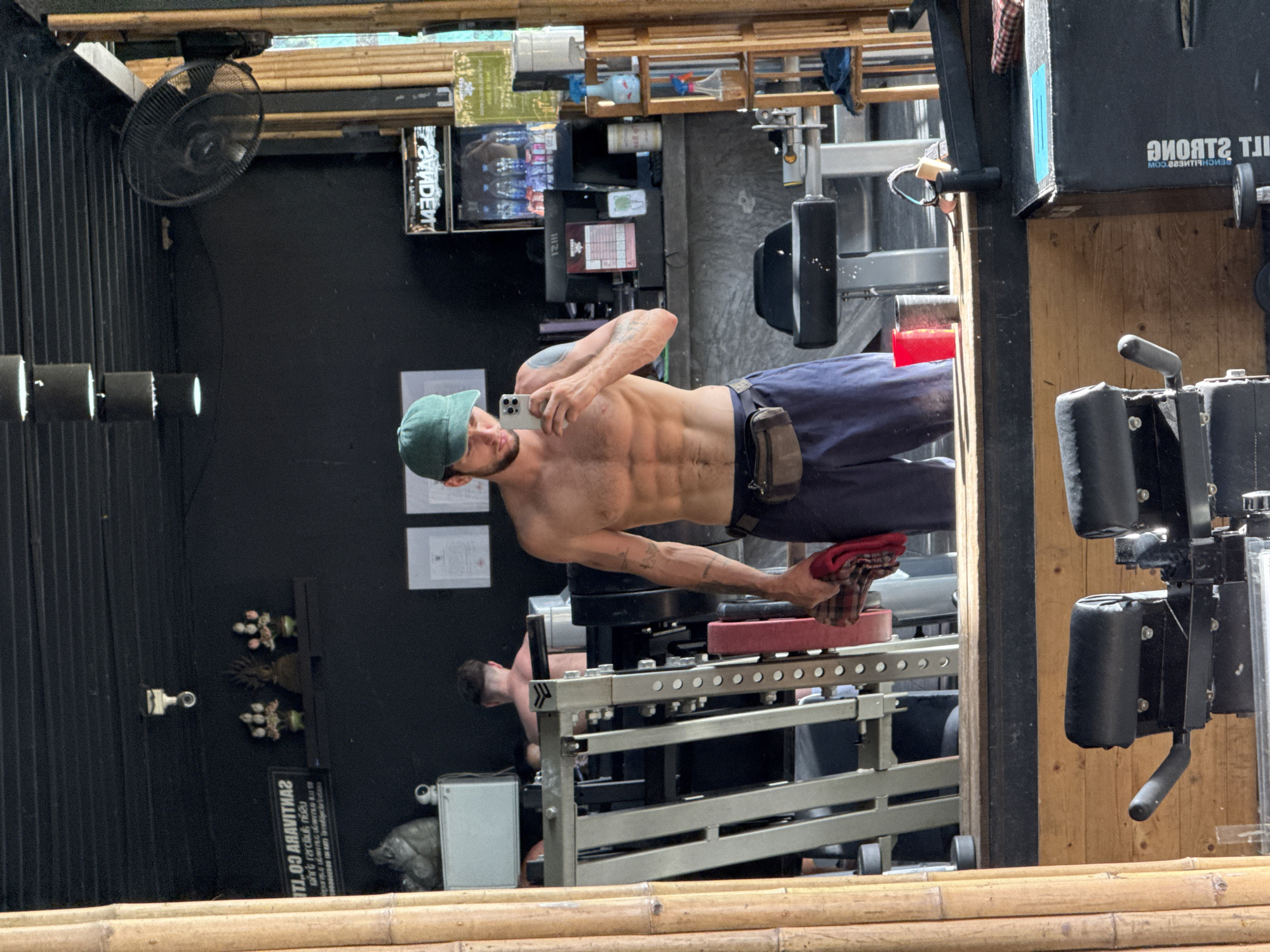 A mirror reflects a man in a gym in Baan Sri Thanu, Thailand. The man, wearing a green cap and blue pants, is shirtless and taking a selfie with his phone. The mirror shows a gym setting with weightlifting equipment and other gym-goers. The gym has a black wall, a bamboo ceiling, and miscellaneous objects on shelves.