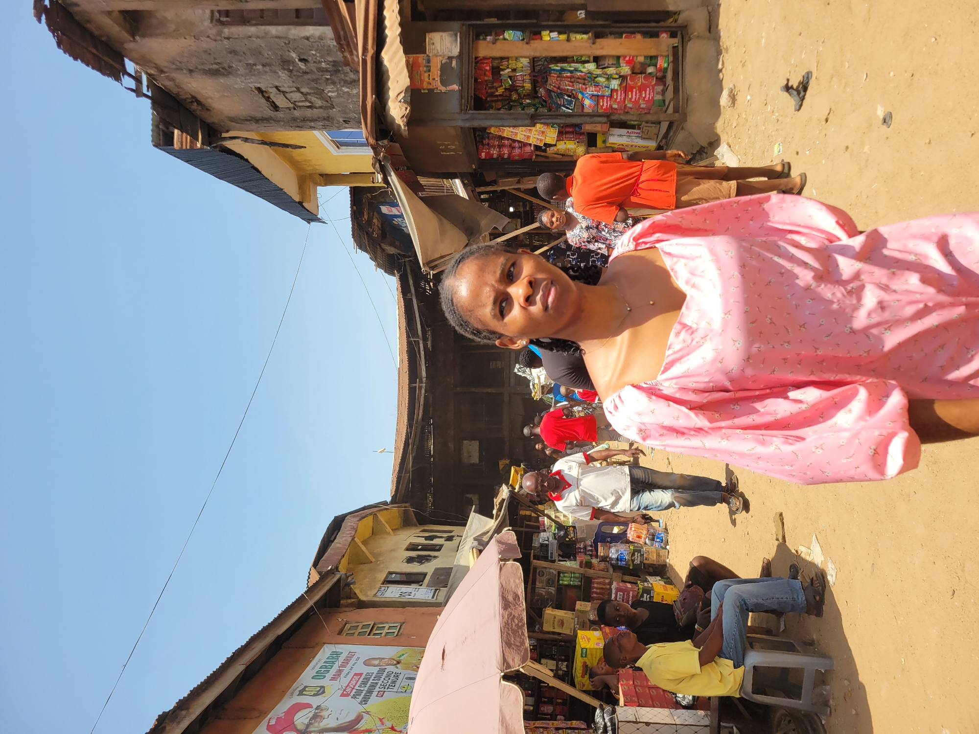 This image captures a vibrant street scene, likely a market or commercial area, in Onitsha, Nigeria. The bright, clear blue sky indicates it is daytime, under sunny weather conditions.
In the foreground, a woman wearing a light pink dress with puffy sleeves and a delicate necklace looks directly at the camera with a neutral expression. Her hair is braided. Behind her and to her left, other individuals are visible, including one in an orange top and another in a black and white patterned shirt, engaged in the market activity.
The street is bustling with people and small businesses. Several stalls and shops line the unpaved, dusty ground, displaying various packaged goods like snacks, drinks, and food items. One shop directly behind the foreground woman is densely packed with products on shelves. Further into the scene, a man in a white and red polo shirt and jeans stands, while two other men are seated on plastic stools near another stall—one in a yellow t-shirt and jeans, and another in a black t-shirt. A large, light pink umbrella provides shade over a section of a stall.
The architecture consists of old, multi-story buildings with corrugated metal roofs, some showing signs of wear. A thin black power line stretches across the upper sky. A notable detail is a large poster affixed to a building on the left side, clearly displaying the text "OGBARU MAIN MARKET," along with smaller text mentioning "PROUD SPONSORS OF BRONXUMA SQUASH 2ND SECOND TOURNAMENT," "ZANO KHAKI," and "CRUNCHIUM DRINKS," featuring a cartoon character. The number "105" is faintly visible on one of the buildings. The scene conveys the lively, everyday atmosphere of an active urban market.