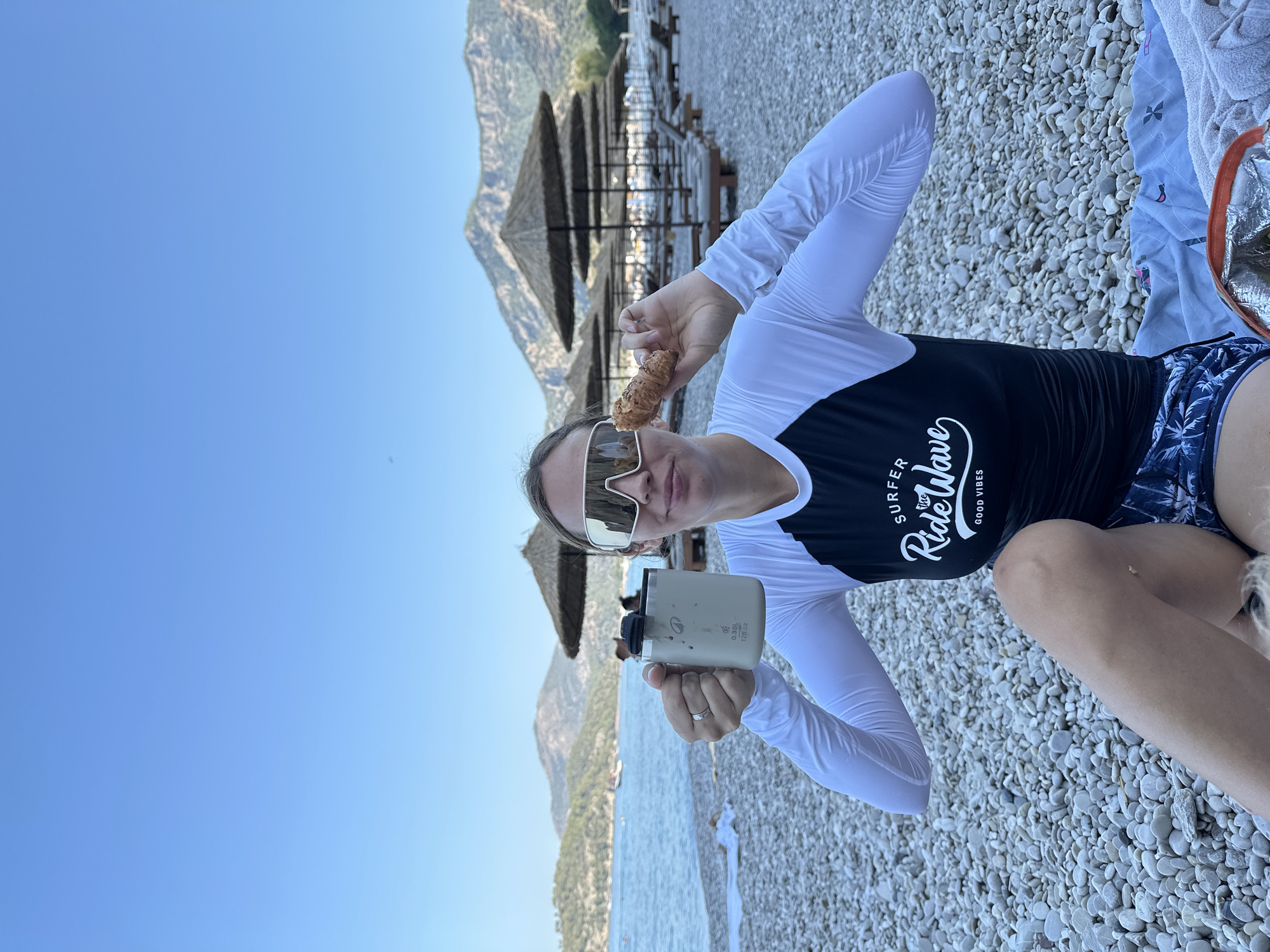 A woman sits on a pebble beach in Fethiye, Turkey, holding a mug in her left hand and a pastry in her right. She wears a long-sleeved rash guard with a black and white design, sunglasses, and blue patterned shorts. The beach has rows of thatched umbrellas and lounge chairs. In the background, the sea meets a hilly, green landscape under a clear blue sky.