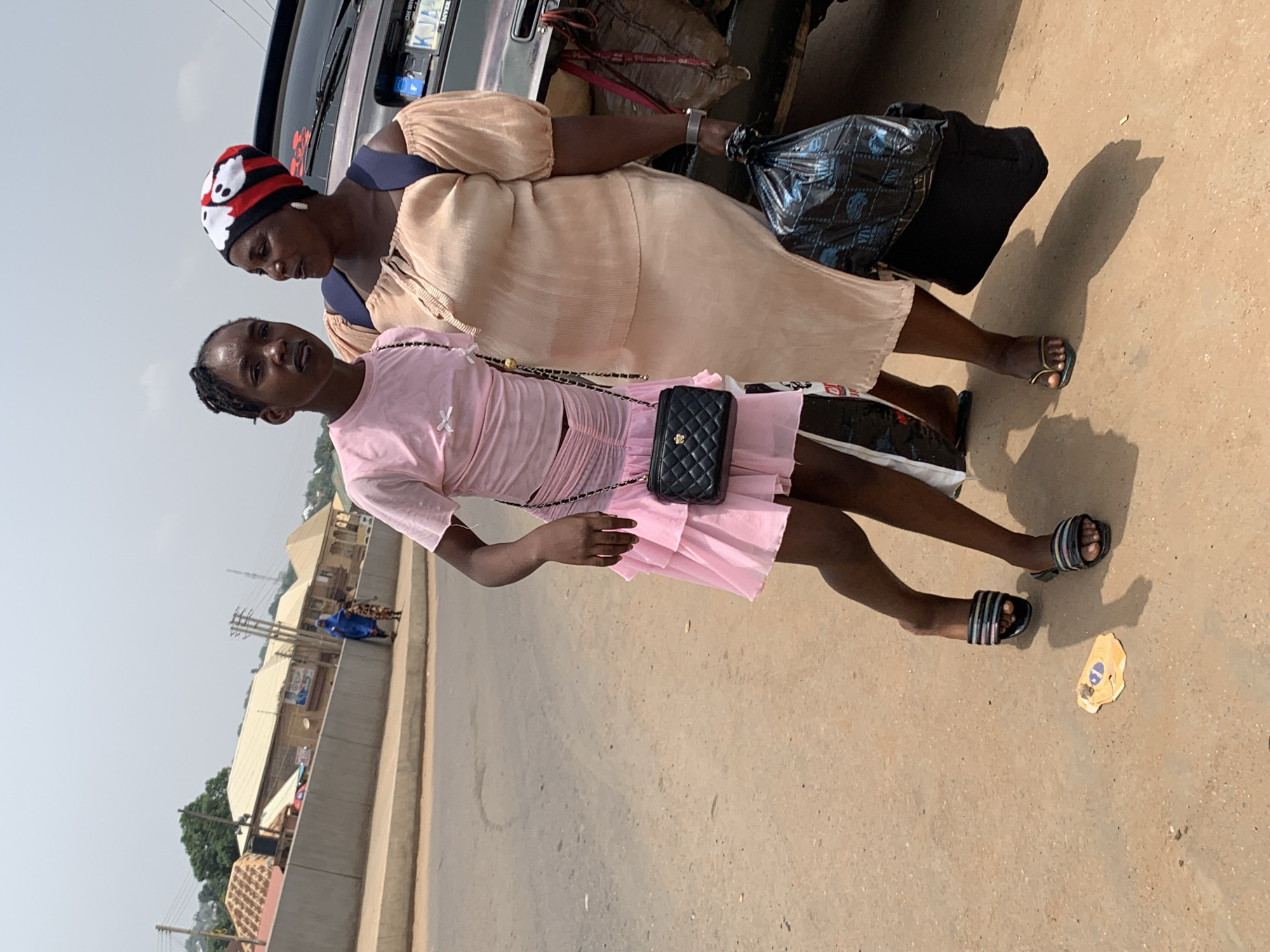 Two women stand on a paved surface alongside a road. The younger woman is wearing a short-sleeved pink dress and black sandals, carrying a small black quilted purse on a chain. The older woman is wearing a beige dress and sandals, with a red and white cap. She is carrying a large black plastic bag. A vehicle is visible behind them. In the background, buildings and trees are visible under a hazy sky. Given the clues and visual characteristics, the location is plausibly Akwanga, Nigeria.