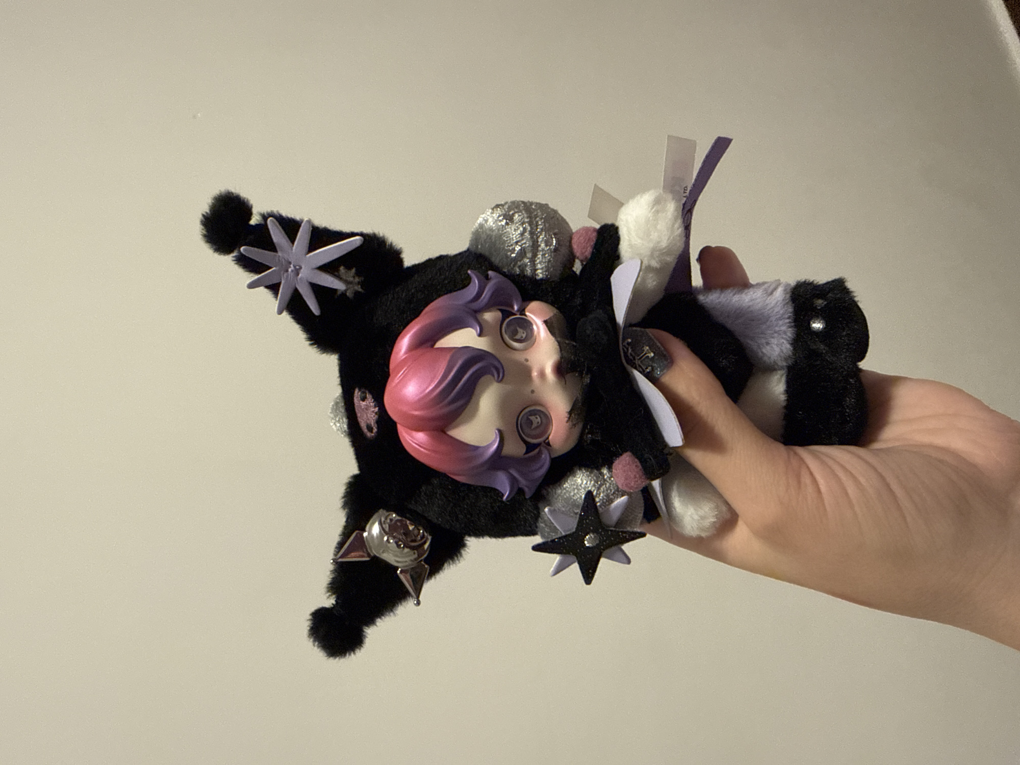 A person holds a small, dark, fluffy toy with a plastic doll face. The toy features star-shaped ornaments and other embellishments. The background is a plain, light-colored wall. The location is not discernible from the image.