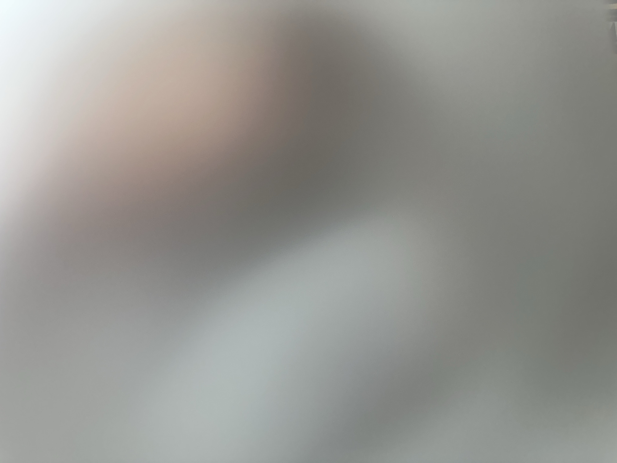 The image is an abstract, out-of-focus depiction of a surface with subtle color variations. The dominant tones are shades of gray, white, and a faint hint of peach or pink. There are no discernible subjects such as people or objects, nor is there a clear scene or setting. The image appears to be a photograph of a textured or diffused surface, possibly fabric, paper, or a wall, with the lighting creating soft gradients and highlights. The abstract nature of the image prevents the identification of any specific activity or event taking place. There are no visible text elements. Given the lack of concrete details, it is impossible to determine the time of day, weather conditions, specific location cues within Belgrade, Serbia, or any emotions conveyed. The overall impression is one of soft light and diffused texture.