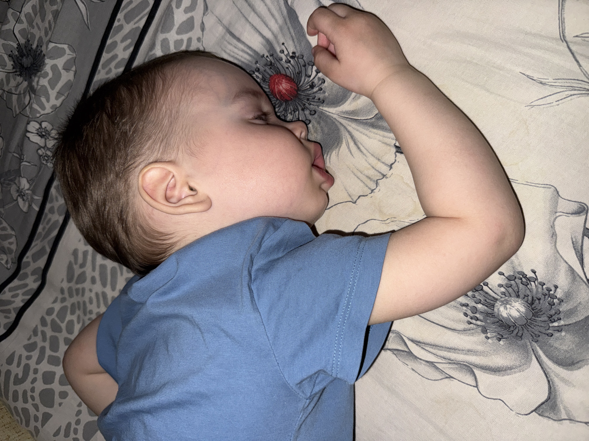 A baby is lying on their side on a bed. The baby has light skin, brown hair, and is wearing a blue t-shirt. Their mouth is slightly open, and their right arm is raised with their hand near a floral design on the bedspread. The bedspread has a floral pattern in shades of gray and white. A location cannot be verified.