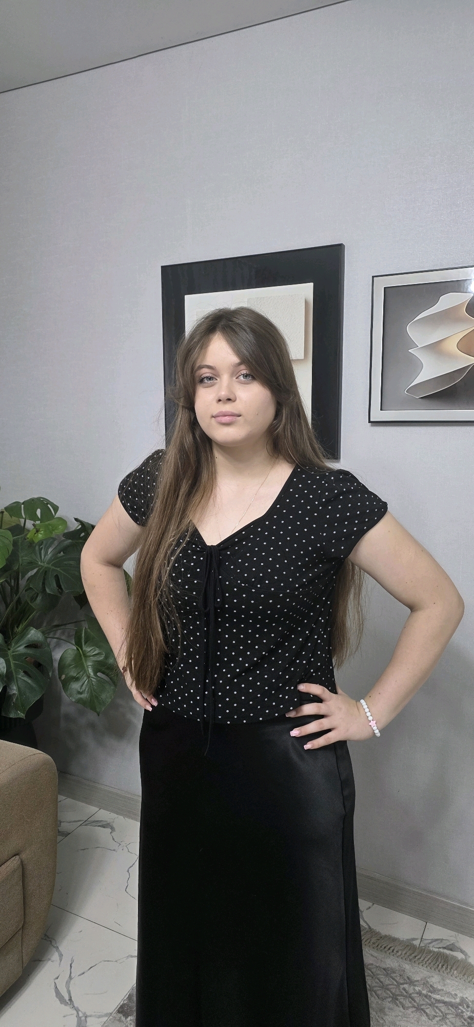 A young woman with long, brown hair and blue eyes is standing in a room, looking at the camera. She is wearing a black polka dot top with a tie at the neckline and a long black skirt. Her hands are on her hips, and she is wearing a bracelet on her right wrist. Behind her, there are two framed pieces of art on the wall, and a large plant is visible to her left. The flooring appears to be tiled. The overall impression is that of a casual indoor portrait. The image does not contain any explicit indicators of its specific location within Almaty, Kazakhstan.