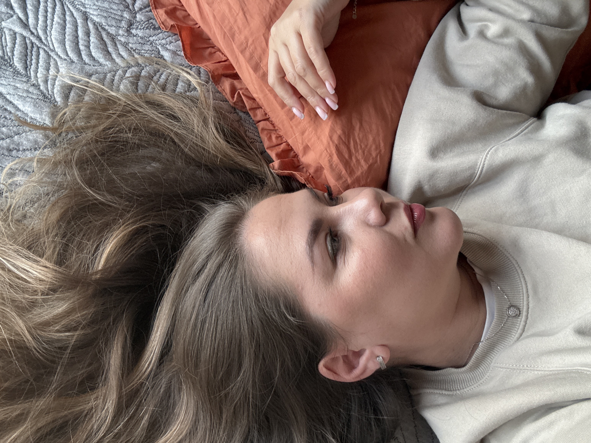 A woman is lying on a gray quilted bedspread with her head on an orange ruffled pillow. She is wearing a beige sweatshirt and a delicate silver necklace with a circular pendant. Her hair is long and light brown, spread out on the bedspread. Her hand rests on the orange pillow, showing light pink and white manicured nails. A small silver earring is visible in her left ear. The scene does not depict any information that relates to the city of Izhevsk, Russia.