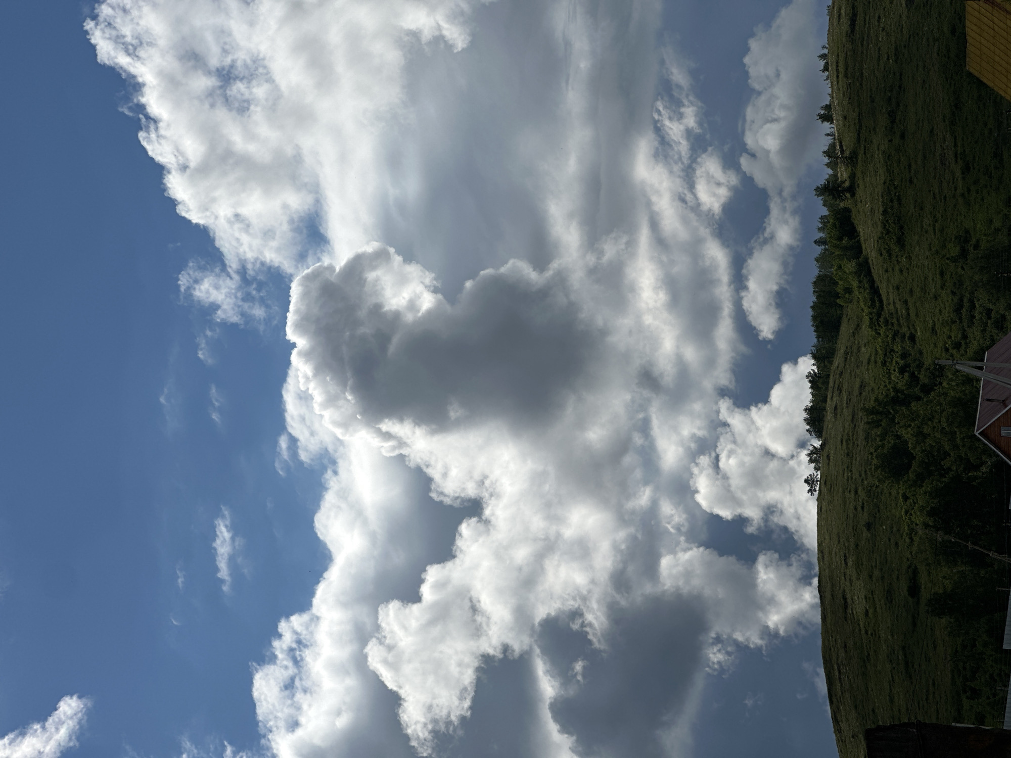 The image shows a view of the sky with various cloud formations, ranging from bright white to dark gray, suggesting different densities and depths. The blue sky is visible in the gaps between the clouds. On the right side of the frame, there is a green, grassy hill with some trees scattered on its slope. The image appears to have been taken with the camera angled upwards towards the sky, with the hill and trees in the periphery. The specific location within the city of Unknown, Unknown is not discernible from the image.