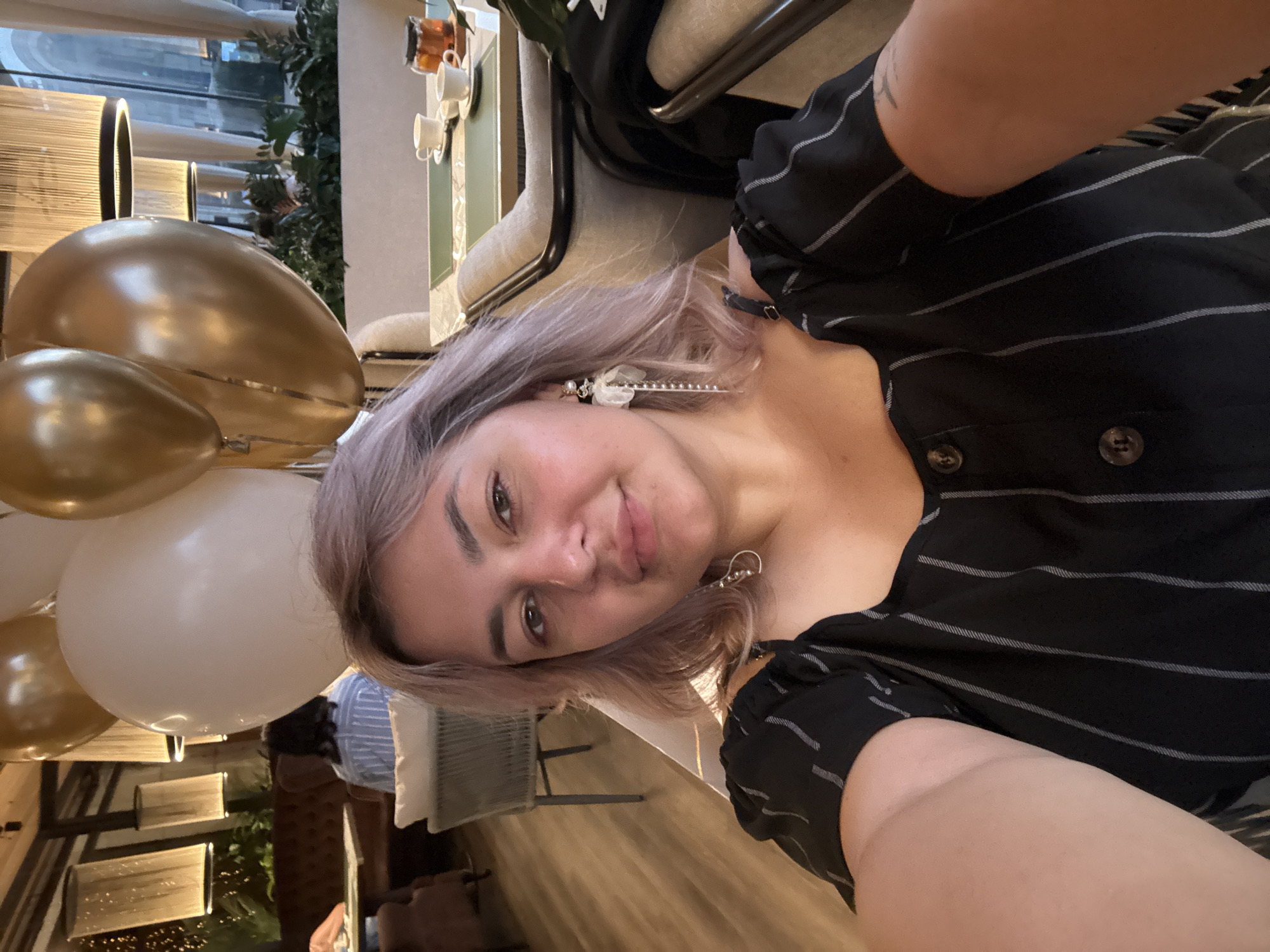 In a well-lit indoor setting, possibly a restaurant or cafe, a woman with light purple hair and light skin takes a selfie. She is wearing a black dress with thin white vertical stripes and has dangly earrings. Behind her, several balloons are visible: two gold balloons and one large white balloon. The background shows glimpses of other patrons, dining tables with teacups, and decorative lamps. The flooring is wooden planks, and there are upholstered chairs and seating areas.