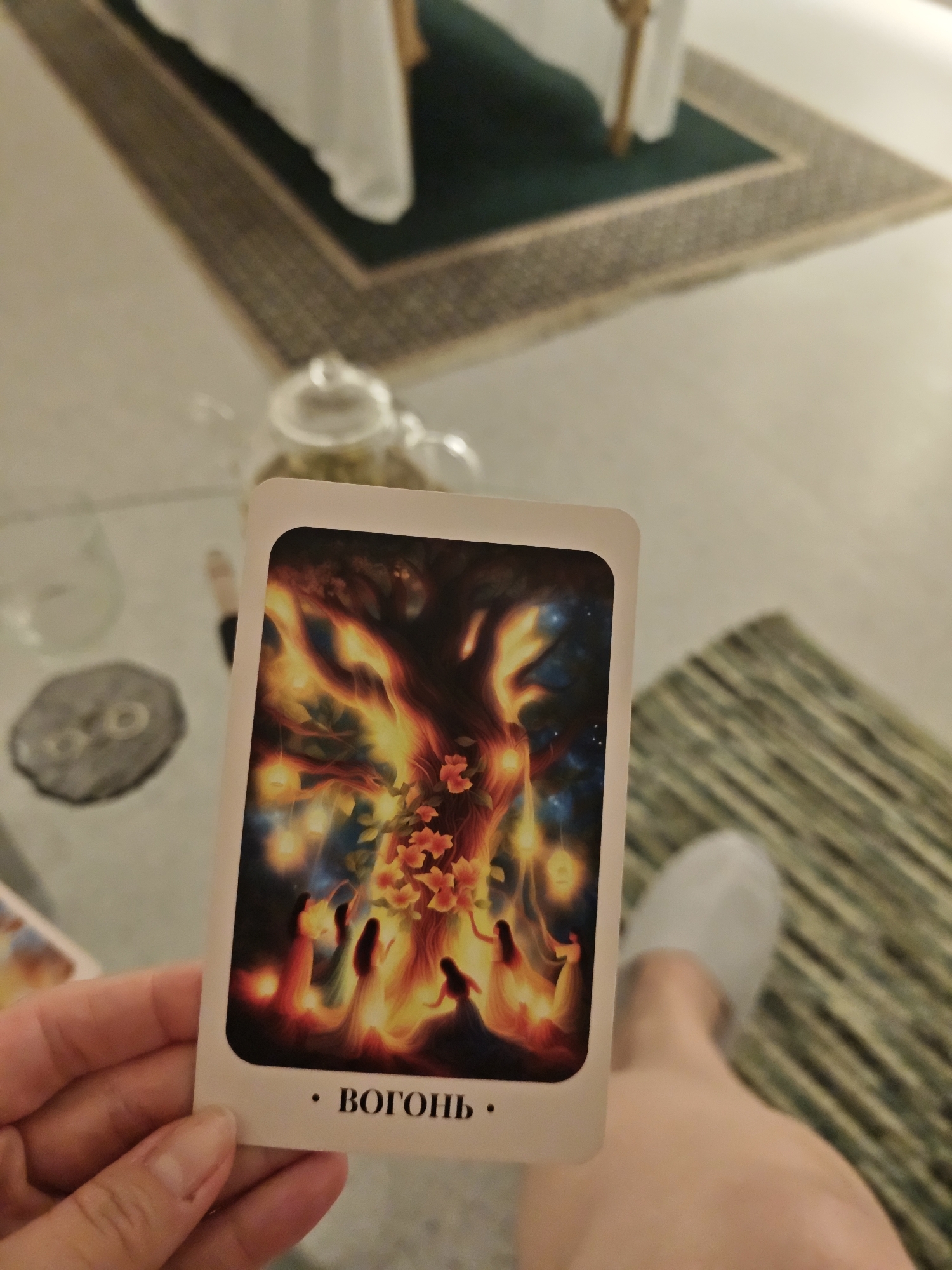 A person is holding a tarot card with an illustration of a fiery tree and figures, with the word "ВОГОНЬ" (Fire) printed on the bottom. The background shows a glass table with a teapot, a decorative object, and a foot with a white slipper. There is a rug on the floor, and furniture is visible in the background. There is no information available to determine that the photo was taken in the city of Vinnytsia, Ukraine.