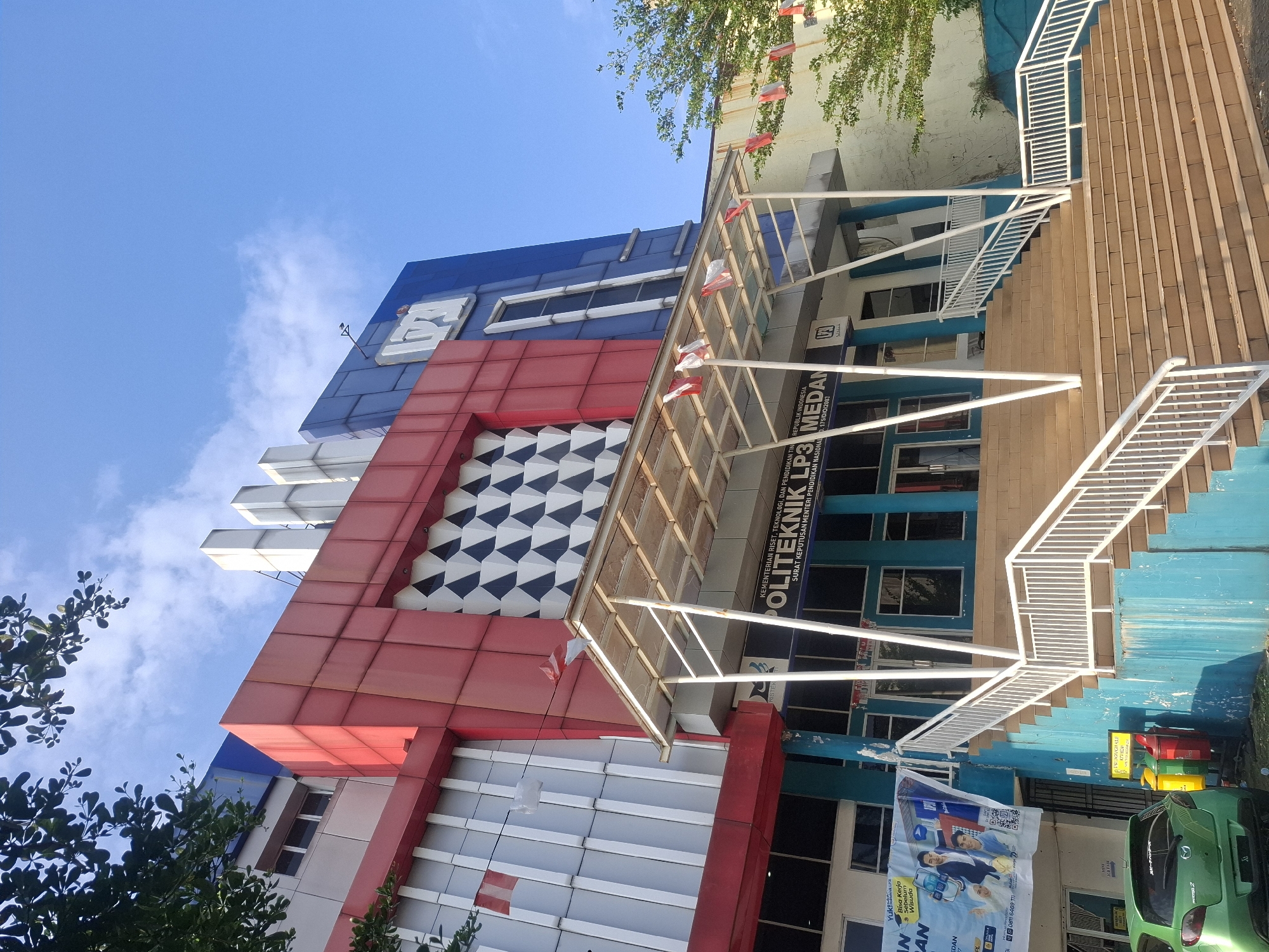 This is a low-angle, medium shot of the front of a multi-story building in Medan, Indonesia. The building has a modern, angular design with sections of red, blue, and white facade. The lower part of the building is painted a bright teal. A wide set of stairs with white railings leads up to the main entrance, which is covered by a white awning. Several small Indonesian flags are strung across the awning. Above the entrance, the words "POLITEKNIK LP3 MEDAN" are visible in large, white letters against a blue background. The sky is clear blue with a few wispy clouds. Trees with green foliage are visible on the left and right sides of the frame. In the lower left corner, a bright green hatchback car is parked. There are also colorful trash receptacles near the base of the stairs. The overall scene suggests a clear, sunny day at an educational institution.