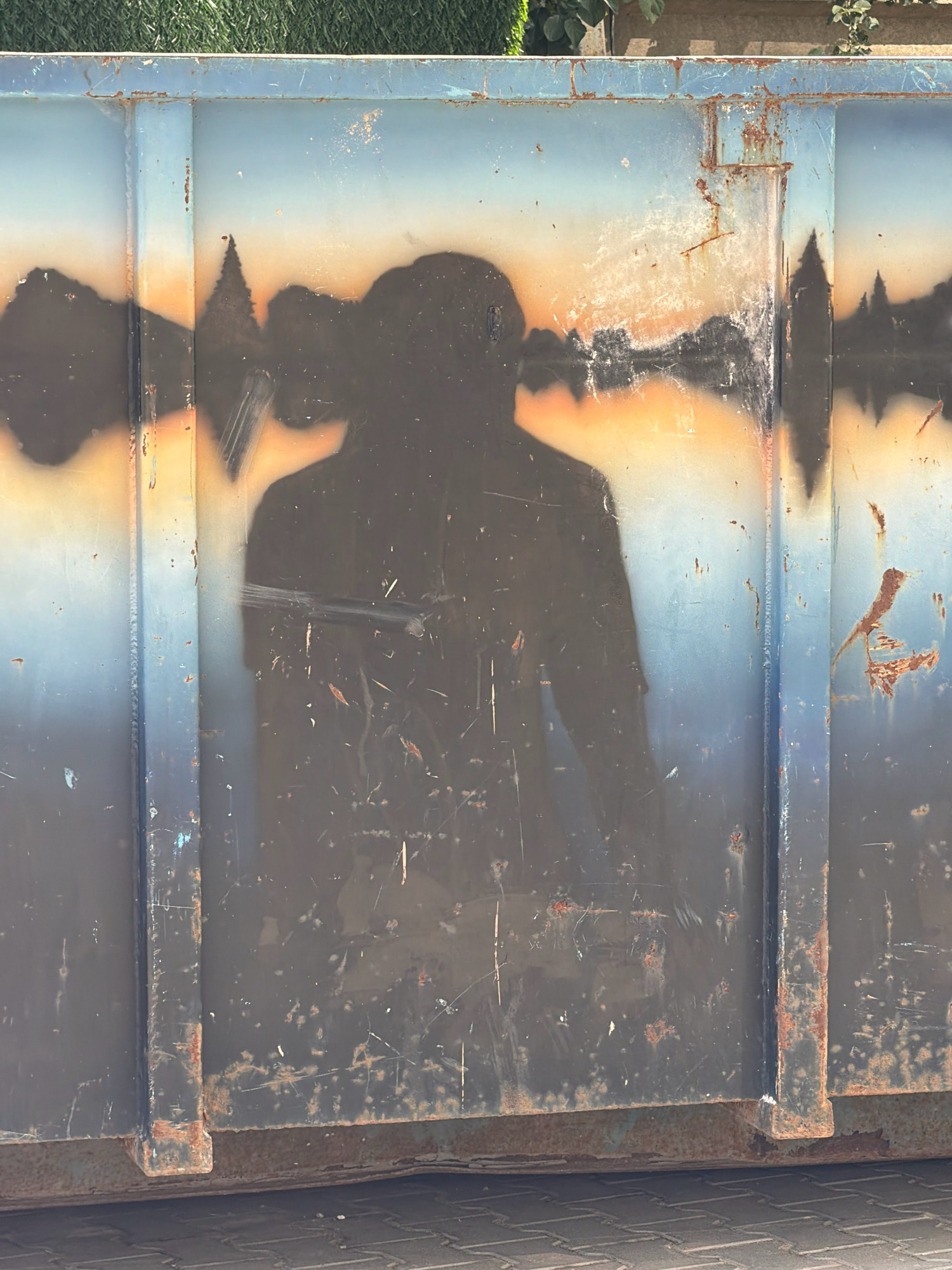 This is a close-up, eye-level, outdoor, vertical, full shot of a large, rusty, blue and black skip bin. The skip bin appears to be made of metal and has visible signs of wear and tear, including rust spots and scratches. A silhouette of a person, with arms extended, is spray-painted or stenciled onto the side of the bin, appearing to be standing in front of a sunset landscape. The silhouette is dark brown and merges with the darker sections of the skip bin. Above the silhouette, the skip bin's surface transitions from a dark color to a lighter blue and then to a gradient of orange and yellow, resembling a sky at sunset. There are also abstract dark shapes suggestive of trees or buildings along the horizon line. The top edge of the skip bin is visible and shows more rust. In the background, a blurred green area, possibly grass or foliage, and a brown wall can be seen. The skip bin is positioned on a grey paved surface, possibly a sidewalk or road. The scene is lit by daylight. There is no clear indication of the city of Kyiv, Ukraine, or any specific location within it.
