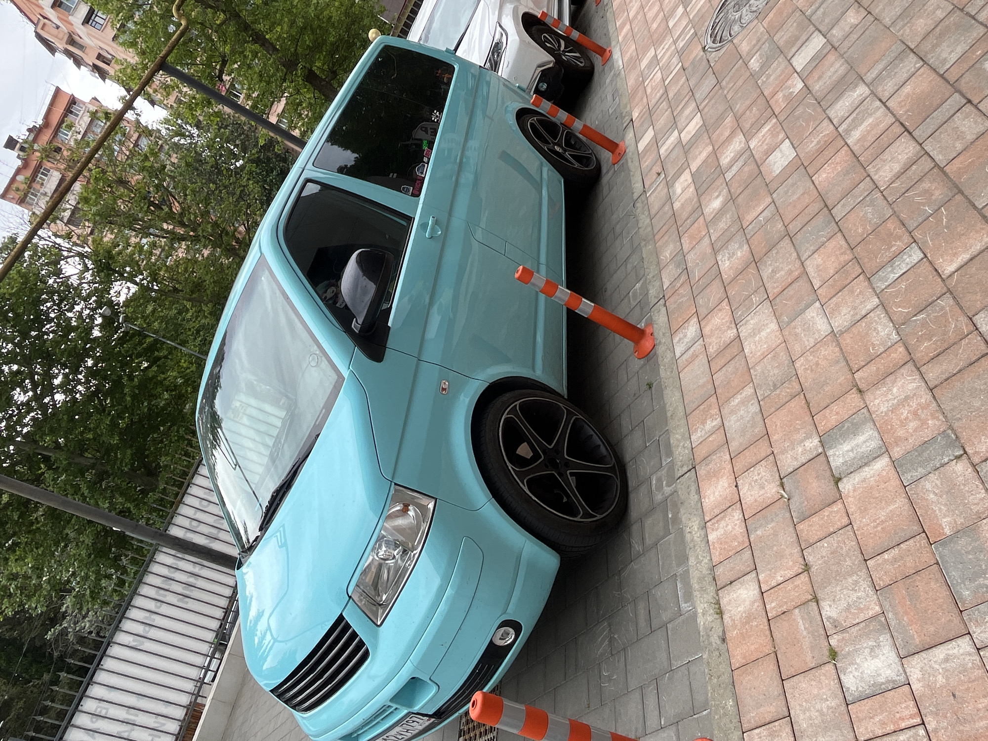 A turquoise Volkswagen T5 van is parked on a sidewalk in Sochi, Russia. The van has tinted windows and black rims. Orange and white traffic cones are placed along the side of the van. In the background, there are apartment buildings and trees. The ground is paved with bricks.