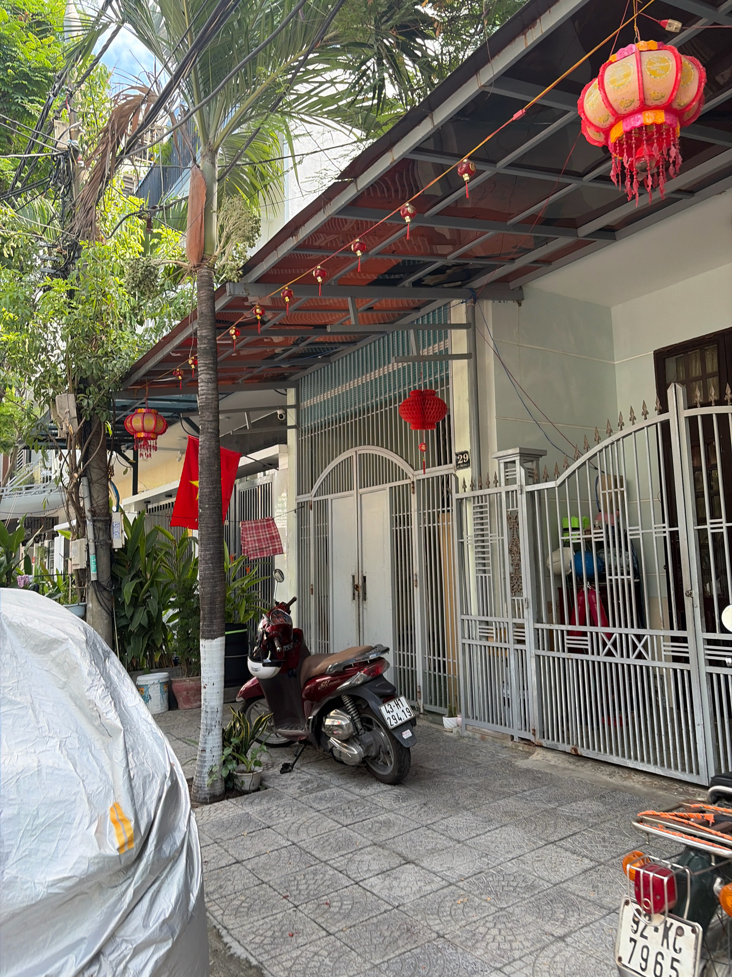 The image depicts the exterior of a house on a street in Đà Nẵng, Vietnam. In the foreground, a motorcycle is parked on a tiled walkway. The house has a metal awning covered with red tiles, decorated with hanging lanterns. A red Vietnamese flag is visible, as well as potted plants and trees. The scene appears to be in a residential area, with other houses and power lines visible in the background. The overall impression is one of a typical street view in a Vietnamese city.