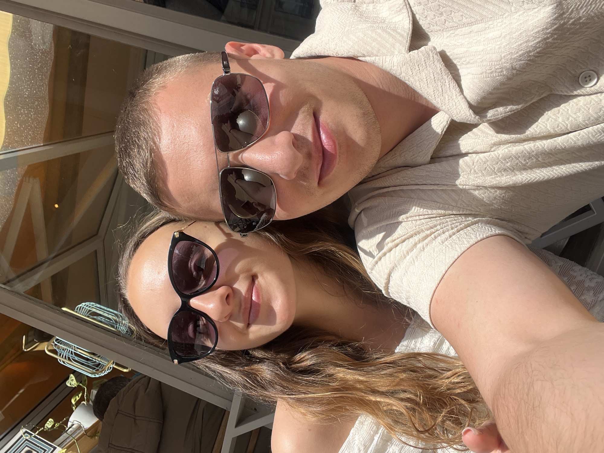 This sunlit selfie captures a man and a woman, likely a couple, in what appears to be an outdoor setting, possibly a cafe terrace or balcony in Paris, France. Both subjects are smiling subtly and wearing sunglasses, indicating bright daylight.
The man is positioned at the top of the frame, facing the camera. He has short brown hair and light stubble on his face. He is wearing a light-colored, possibly cream or beige, textured button-up shirt with a subtle geometric pattern. His aviator-style sunglasses have a silver frame, and their lenses reflect window frames and an indoor space.
Below him, the woman looks up at the camera with a soft smile. She has long, wavy brown hair with lighter highlights, which flows over her shoulder. She is wearing a light-colored, possibly white or cream, top. Her dark-framed cat-eye sunglasses clearly reflect window panes, a light blue wire basket, and a potted plant, strongly suggesting an outdoor seating area looking into an establishment.
The strong, warm sunlight illuminates their faces and clothes, casting defined shadows and highlighting the texture of their skin, hair, and garments. The overall impression is one of relaxation and happiness on a bright, sunny day, characteristic of enjoying a leisurely moment in a city like Paris. No text is visible in the image.