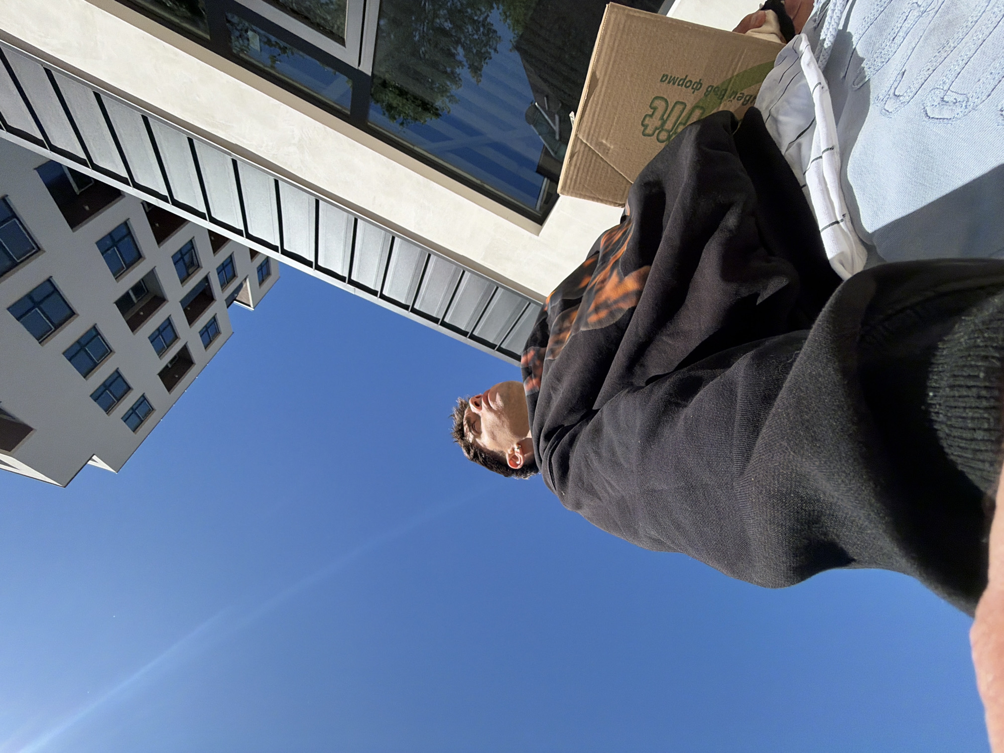The image is taken from a low angle, looking up at a person carrying items under a bright blue sky. In the background, there are buildings with modern architectural designs, suggesting an urban environment. The person appears to be wearing a black and orange hooded sweatshirt, a light blue garment, and a white striped piece of clothing. They are also carrying a cardboard box with lettering in what appears to be Bulgarian. Due to the Bulgarian text on the box, it can be reasonably inferred that the location is likely Sofia, Bulgaria.