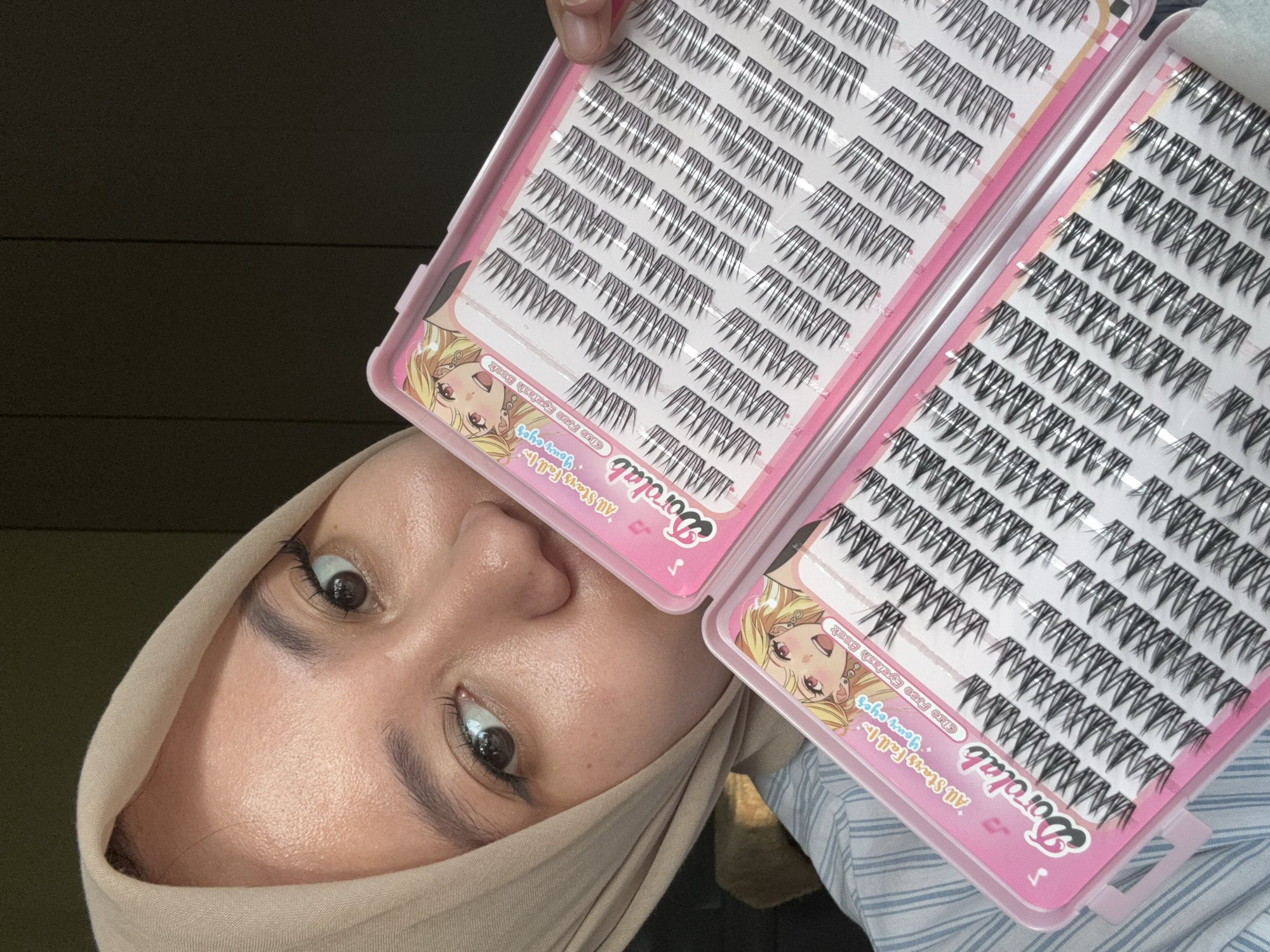 A woman wearing a hijab is holding two open pink cases of Dorolab false eyelashes. The cases contain rows of individual lashes. The location is in Gadingrejo, Indonesia.