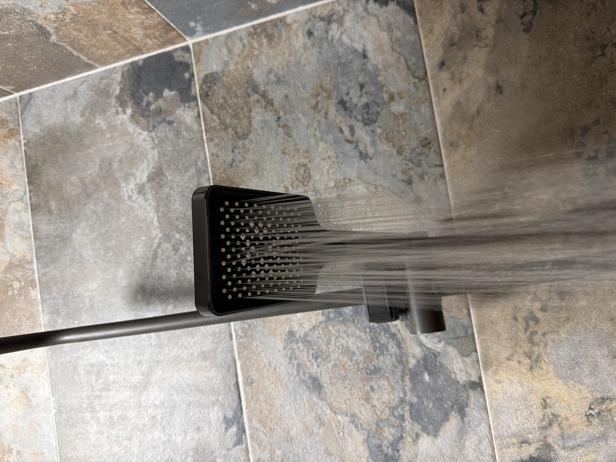 A close-up, eye-level shot shows a dark, rectangular shower head with water spraying from its numerous nozzles. The shower head is attached to a dark metal pipe that extends upwards. The background consists of a tiled wall with large, irregular tiles in shades of grey, brown, and blue, resembling slate. The tiles have a textured, naturalistic appearance. The lighting appears to be indoor, possibly from a bathroom fixture. There are no people visible in the image. The scene is clean and modern, consistent with a bathroom setting. The location in Sofia, Bulgaria is not discernible from the visual information provided.