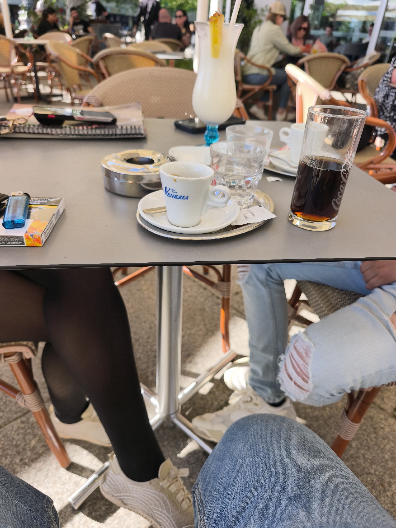 The image presents a close-up view of a table setting in what appears to be an outdoor cafe, with details of the people present. The table is covered with an array of objects: cups, glasses, an ashtray, a cigarette pack, a lighter, and a drink with a straw and pineapple garnish. One glass is filled with a dark liquid, possibly cola. In the background, there are more tables and chairs, indicating an outdoor dining area. People are seated at these tables, enjoying what looks like a leisurely cafe atmosphere. The lower portion of the frame captures the legs of two individuals seated at the table. One person is wearing black tights and white sneakers, while the other is in light-wash ripped jeans and white sneakers. Their positions suggest they are engaged in a conversation or activity together. The details do not include location, only the location of "Unknown, Unknown" which is unspecified.