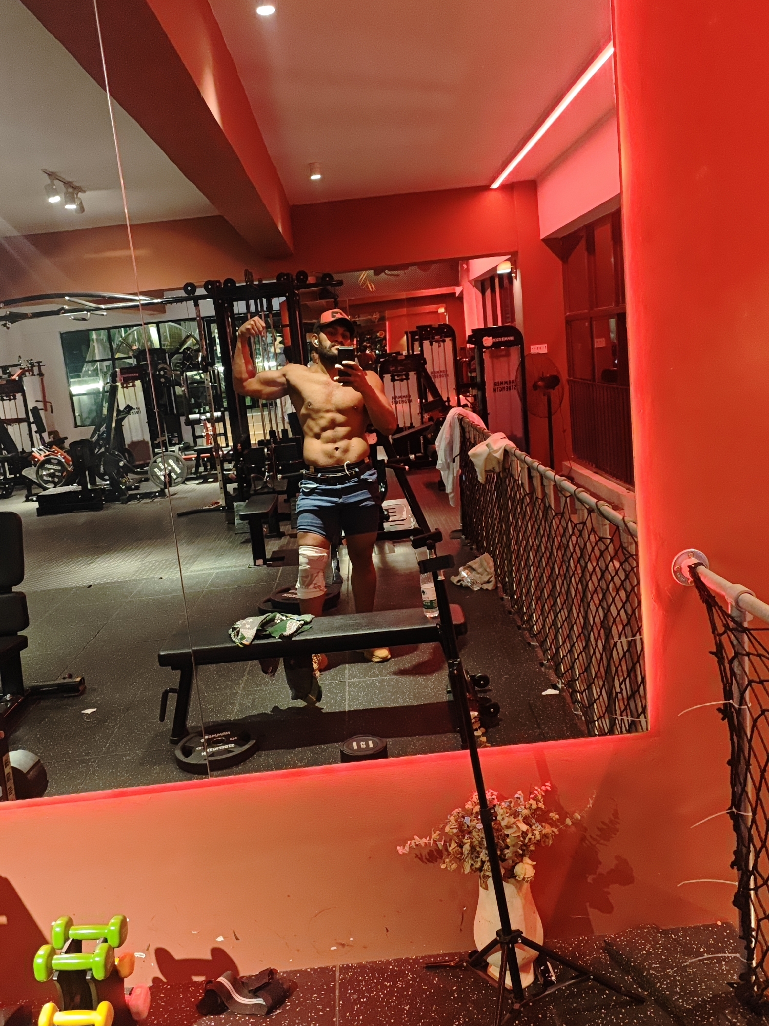 The image displays an indoor gym facility in the Chongqing High-tech Zone, China. The environment is illuminated by overhead white lights and prominent red LED strip lighting along the ceiling and walls, casting a strong red hue across the space. The walls are a reddish-orange color, and the floor is a dark, speckled gym matting.
A shirtless adult male with a muscular build, wearing blue denim shorts and a red baseball cap, is captured in a large mirror taking a selfie with a black smartphone held in his right hand. He is flexing his left bicep, and his left knee is wrapped in a white bandage or brace.
In the background and reflected in the mirror, various gym equipment is visible, including weight machines (some with "HAMMER STRENGTH" branding), several weight benches, and weight plates. In the foreground, on the floor to the left, green and yellow dumbbells are present. On a bench in the reflection, a green and white patterned cloth item is placed, along with a water bottle. To the right, a black net fence runs alongside a raised section, draped with a white towel. A white vase containing dried floral arrangements stands on the floor near the mirror, accompanied by a black tripod. A large red pillar is visible on the far right of the frame.