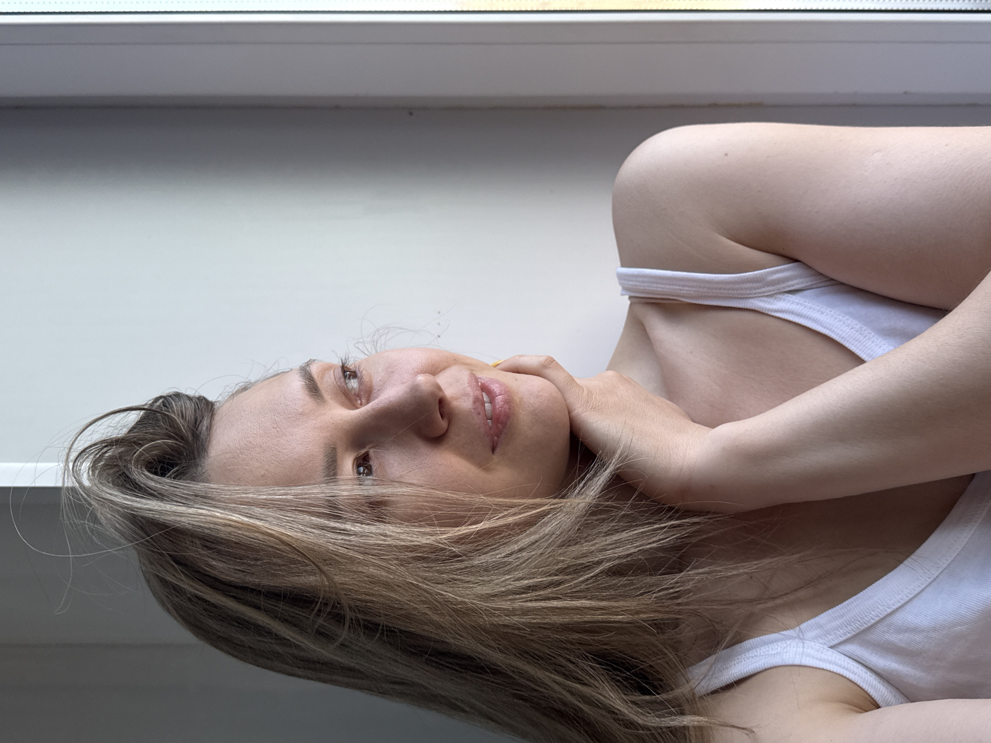 The image, taken in Chaikovsky, Russia, shows a young adult female reclining indoors, likely positioned beside a window. She has light skin and long, wavy hair with shades of brown and blonde, which is spread out around her head and shoulders. She is wearing a simple white tank top or camisole. Her head is slightly tilted, and she gazes upwards and to her left with a gentle, thoughtful, or serene expression, her lips slightly parted. Her right hand is casually placed near her neck and chin. The background is a plain, light-colored surface, possibly a wall or window frame, with a window sill visible at the top, indicating the presence of natural light, suggesting it is daytime. The scene evokes a sense of quiet relaxation. No visible text is present in the image.