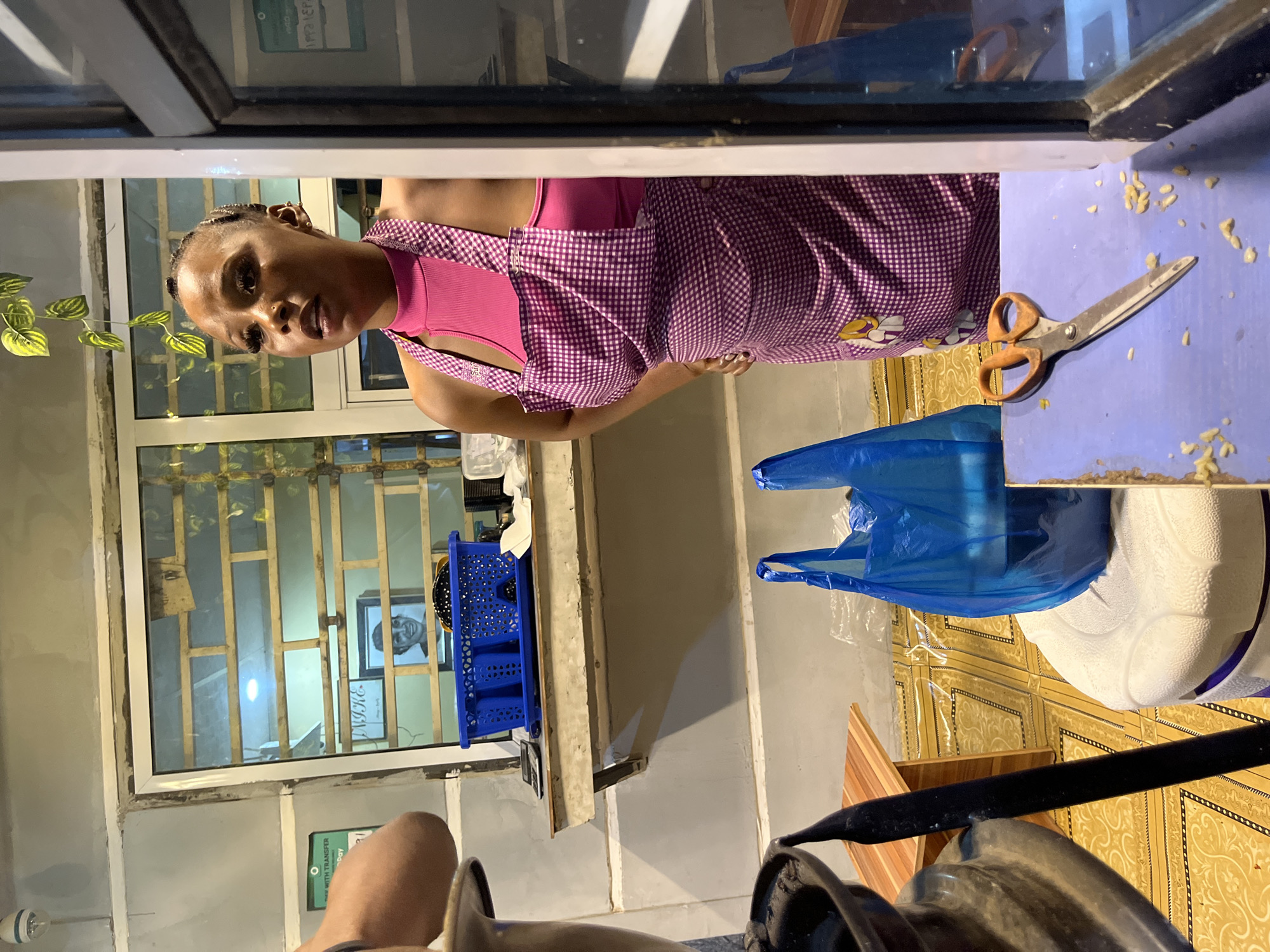 A woman with dark hair and a pink top and checkered apron stands at a blue countertop. On the countertop are a pair of scissors and a blue plastic bag. Behind her is a window with bars and a picture on the wall. The image appears to have been taken from inside a shop or food stand. There is a sign for "WITH TRANSFER today" on the wall. There is a white cooler, and a black bar, possibly belonging to a bicycle. The location is Dushepe, Nigeria.