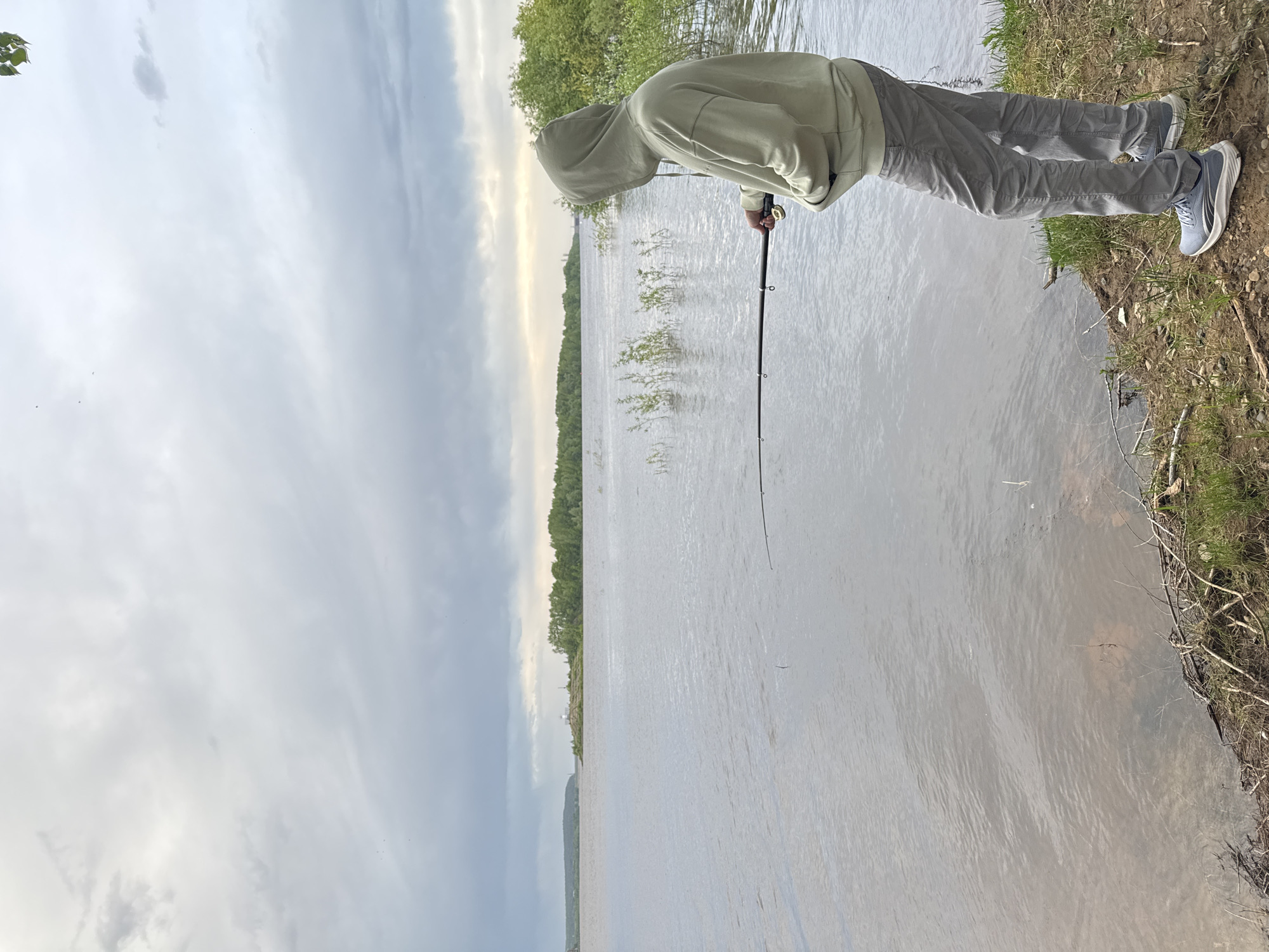 A person wearing a hooded sweatshirt and gray pants stands on a grassy bank at the edge of a body of water, holding a fishing rod. The water extends into the distance, with trees lining the opposite shore. Overcast skies are visible above. The location is Chaikovsky, Russia.