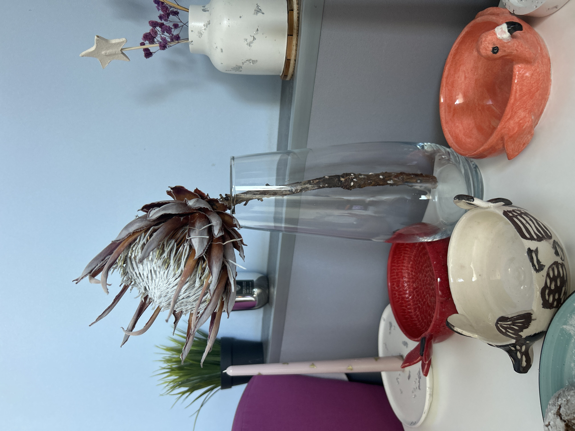 The image depicts a still life arrangement on a white surface, likely a table or shelf, against a light blue wall. In the center, a large, dried Protea flower stands tall in a clear glass vase. To the left of the Protea, a slender pink candle with gold heart patterns is placed on a white plate. Behind the candle, a small artificial green plant in a black pot is visible. Further to the left, a portion of a magenta chair can be seen.
On the shelf above, a white vase with textured detailing holds dried purple flowers and a small star ornament on a stick. To the left of this vase, a metallic cylindrical object is present.
In the foreground, on the white surface, there are several decorative bowls. A vibrant red bowl with a textured surface and small handles is positioned to the left of the flamingo bowl. To its left is a cream-colored ceramic bowl with a bird or animal motif painted in brown. To the right of the bird bowl is an orange ceramic bowl shaped like a flamingo. A corner of a teal plate with a powdered pastry is visible in the bottom left of the frame.
The overall scene is clean and well-lit, suggesting an indoor setting, possibly a home or a retail display. There are no people or actions depicted in the image. This image does not provide any information that would allow for its location to be identified as Orenburg, Russia.