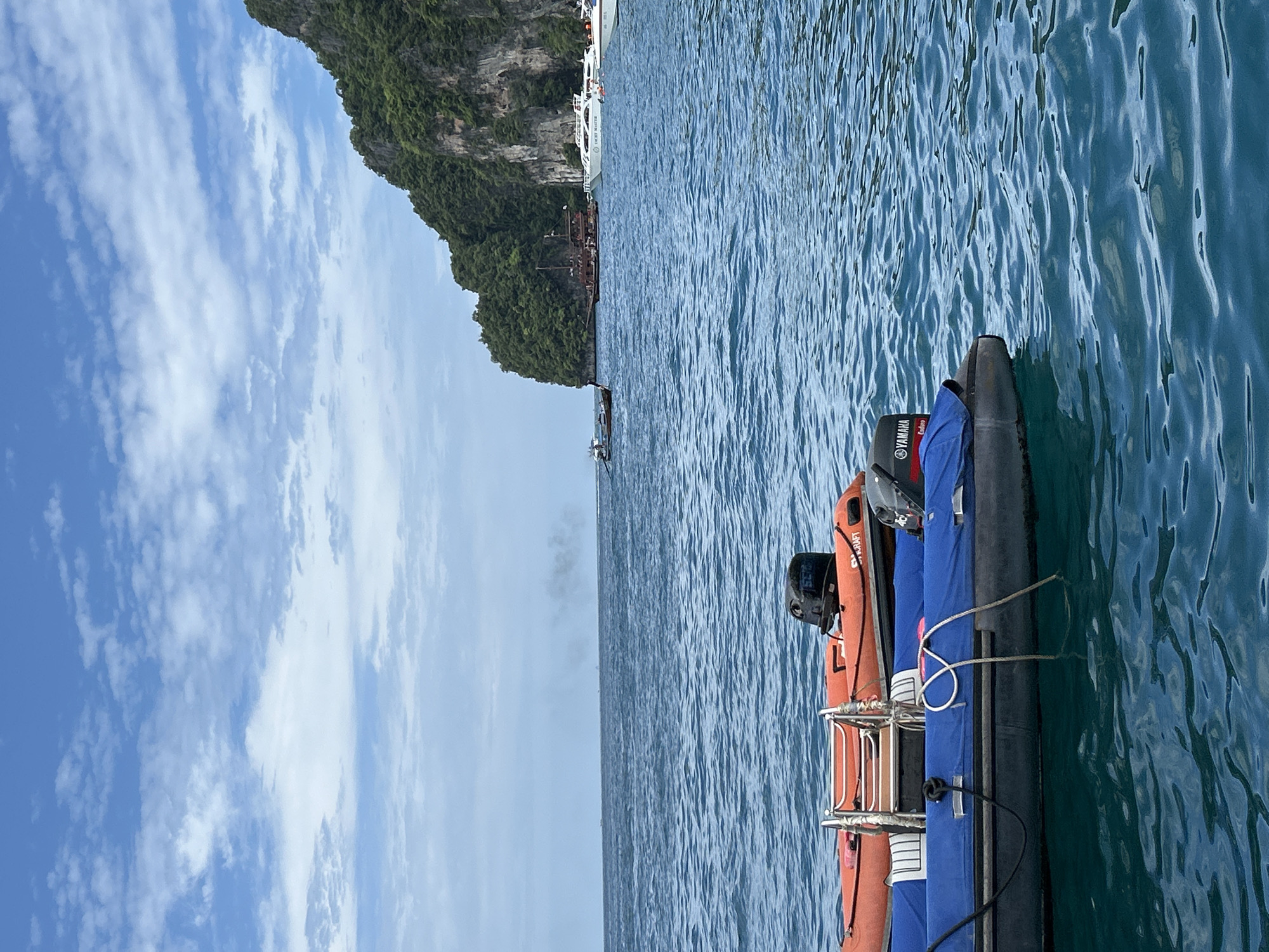 This is a photograph taken from the water, likely from a boat, in Ao Nang, Thailand. In the foreground, two inflatable dinghies are visible, one orange and one blue, each with an outboard motor. The orange dinghy has a metal ladder stowed on it. The blue dinghy is covered with a blue tarp and has a rope tied to it. The water is a deep blue, with moderate waves. In the middle ground, several other boats are anchored or moving in the distance, including what appears to be a larger, older wooden ship and several white speedboats. Beyond the boats, a large, steep, tree-covered limestone cliff rises dramatically from the sea, characteristic of the landscape in Ao Nang. The sky is partly cloudy, with white clouds scattered across a bright blue sky. There are no people clearly visible in the image. The overall scene depicts a typical view of the Krabi coastline, known for its karst formations and numerous islands, often accessed by boat from Ao Nang.