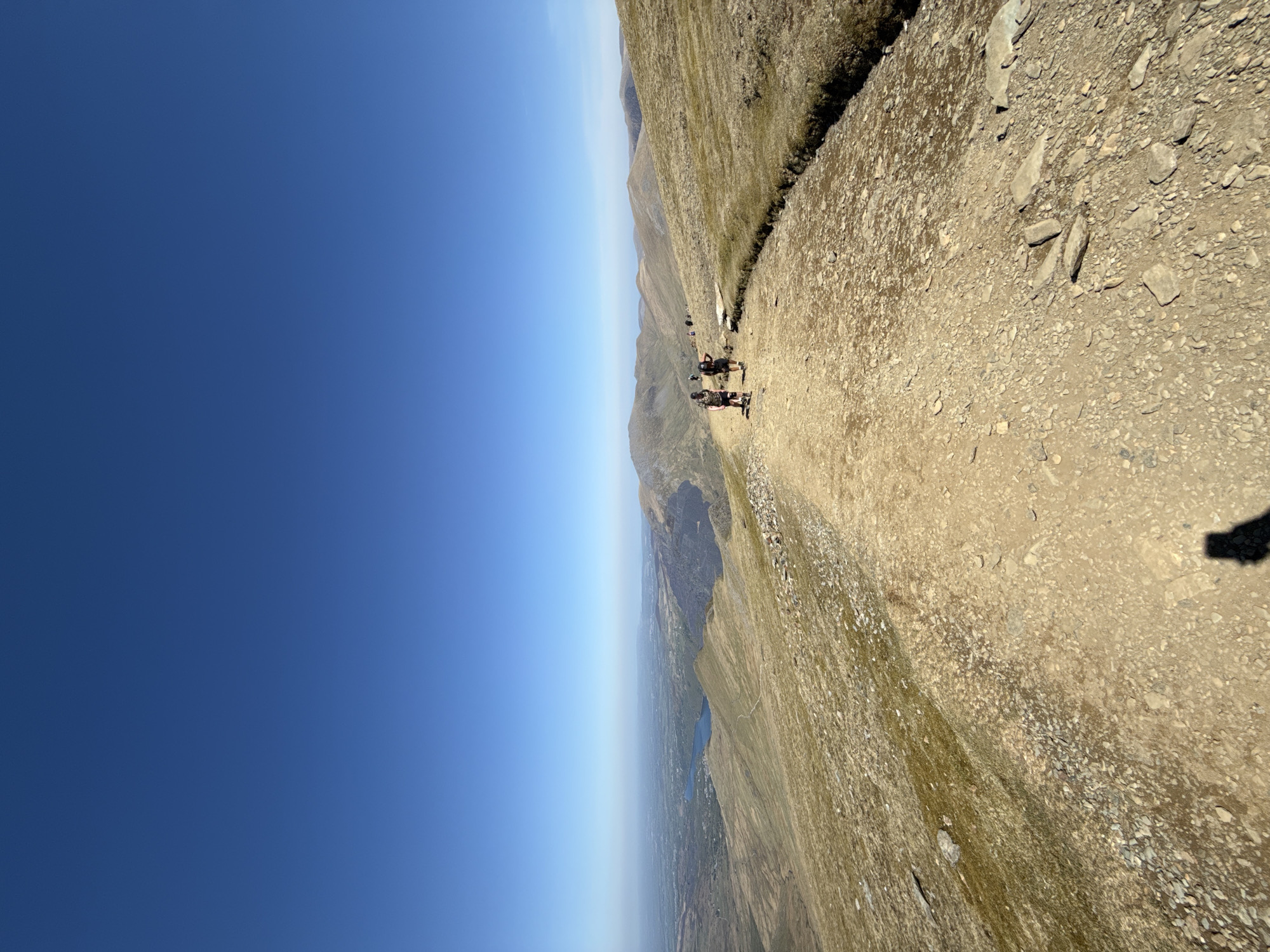 The photograph depicts a mountainous terrain in the vicinity of Llanberis, United Kingdom. The view is predominantly sky and landscape, with a hiking path traversing the side of the mountain.
Several individuals are located on the path, appearing as hikers, though specifics about their clothing or activities are not clearly discernible. The terrain is rocky and barren, with shades of brown and yellow characterizing the ground.
In the distance, a body of water, possibly a lake or reservoir, is visible amid the landscape below. The sky is clear, transitioning from a dark to a light blue hue.