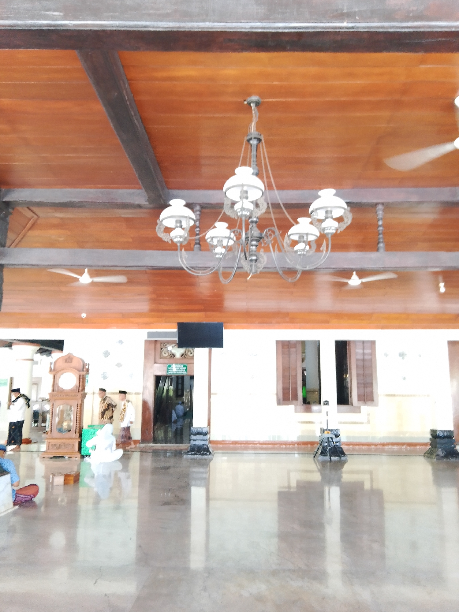 The image captures the interior of what appears to be a mosque in Demak, Indonesia, likely the Grand Mosque of Demak. The ceiling is constructed of polished wooden planks supported by dark brown beams. A traditional ornate chandelier with multiple white lamps hangs centrally. Two white ceiling fans are visible, one on the left and another on the right, with their blades in motion, indicating they are operational.
The wall below the ceiling is painted a light cream color. On the wall, a black rectangular screen or monitor is mounted. To the left of the screen is a tall, antique wooden grandfather clock with a visible clock face. Several individuals are present in the space. Two men in traditional attire, including peci hats, stand near the grandfather clock. Another person in white religious attire is seated or kneeling on the polished floor. Further into the background, a doorway is visible with a green sign indicating a direction, suggesting an exit or another section of the building.
The floor is a highly polished, reflective marble or similar material, mirroring the architectural elements and figures present. In the foreground, to the far left, a person wearing a blue garment is partially visible, seated on a low bench with a patterned cushion. The overall ambiance suggests a place of worship or historical significance, characterized by traditional architecture and religious observances.