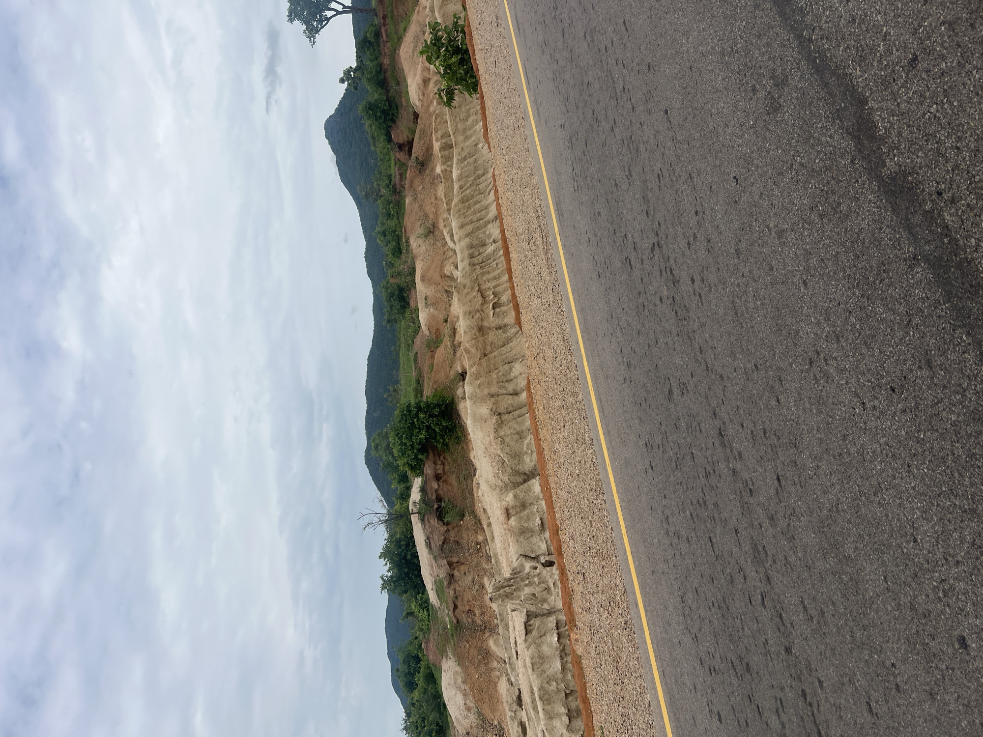 The image shows a road next to a hillside. The road is paved with asphalt and has a yellow line on the left side. The hillside is made of dirt and rock, with some vegetation growing on it. In the background, there are mountains and trees. The sky is cloudy. The image appears to have been taken during the day. There is no text visible in the image. The scene suggests a rural or undeveloped area, possibly in a tropical or subtropical region, given the vegetation and mountainous terrain. The road appears to be recently constructed or maintained, as indicated by the clear yellow line and the absence of significant wear and tear. The cloudy sky suggests it might be a humid or potentially rainy day.