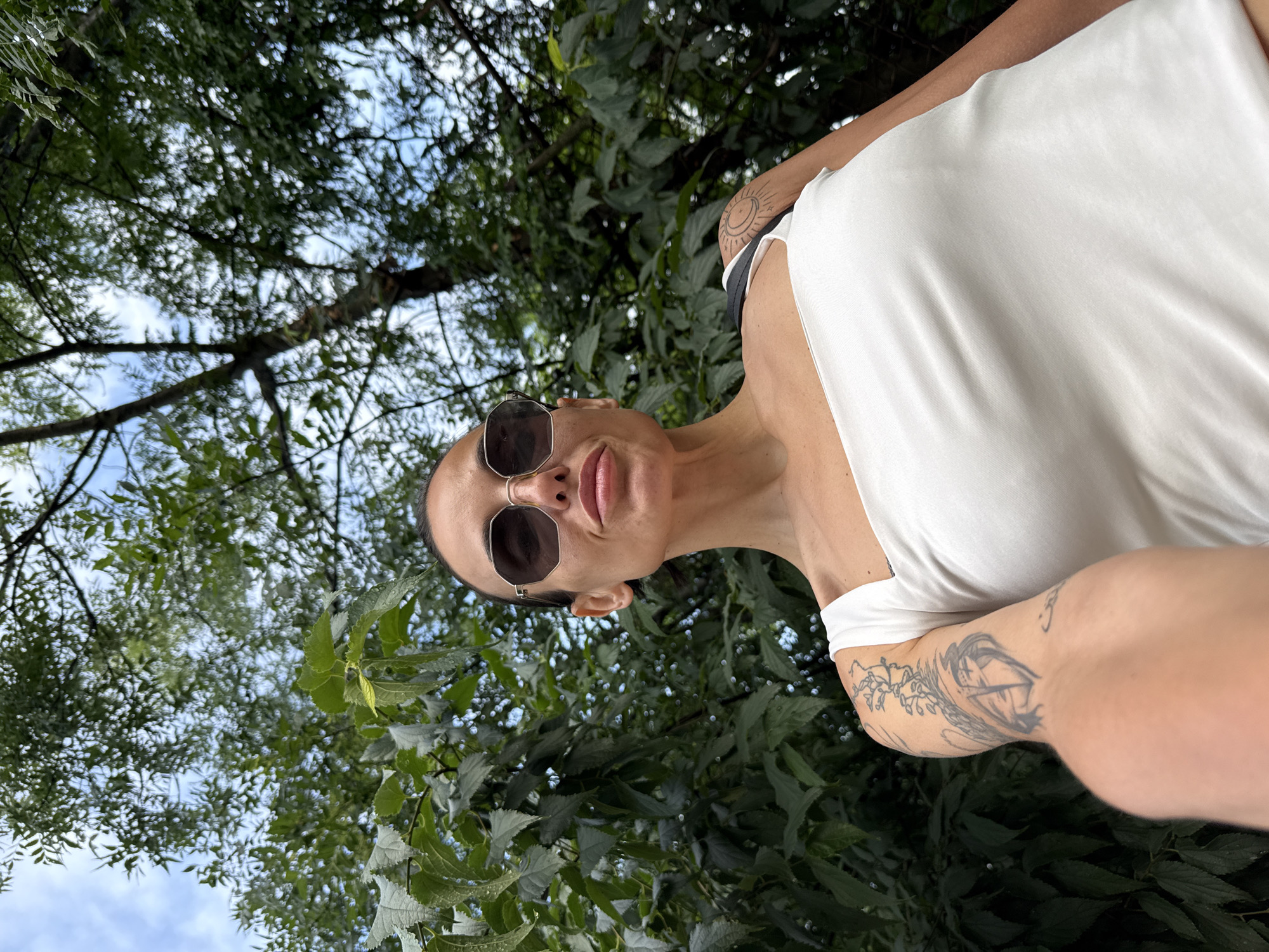 The image is a unique portrait-style shot of a woman, taken outdoors from a low angle and rotated 90 degrees counter-clockwise. The woman's head is positioned in the lower-left part of the frame, angled upwards towards the camera. She wears dark, hexagonal-framed sunglasses, obscuring her eyes, and her lips are slightly parted in a subtle pout. Her hair is pulled back, revealing her ears. She is dressed in a plain white sleeveless or tank top, which highlights her tanned skin and several tattoos. On her upper right arm, a circular tattoo featuring a sun or moon motif is visible. Her left forearm displays a more intricate tattoo with organic, branching patterns.
The background consists of a dense canopy of green tree leaves and branches, creating a lush, natural environment. Through the gaps in the foliage, patches of light blue sky with scattered white clouds are visible, indicating that the photo was taken during the daytime. The perspective suggests the woman is looking up at the camera, which is positioned above her.
The overall scene conveys a relaxed outdoor moment, possibly in a park or garden setting, within the context of Pleven, Bulgaria. There is no visible text in the image.