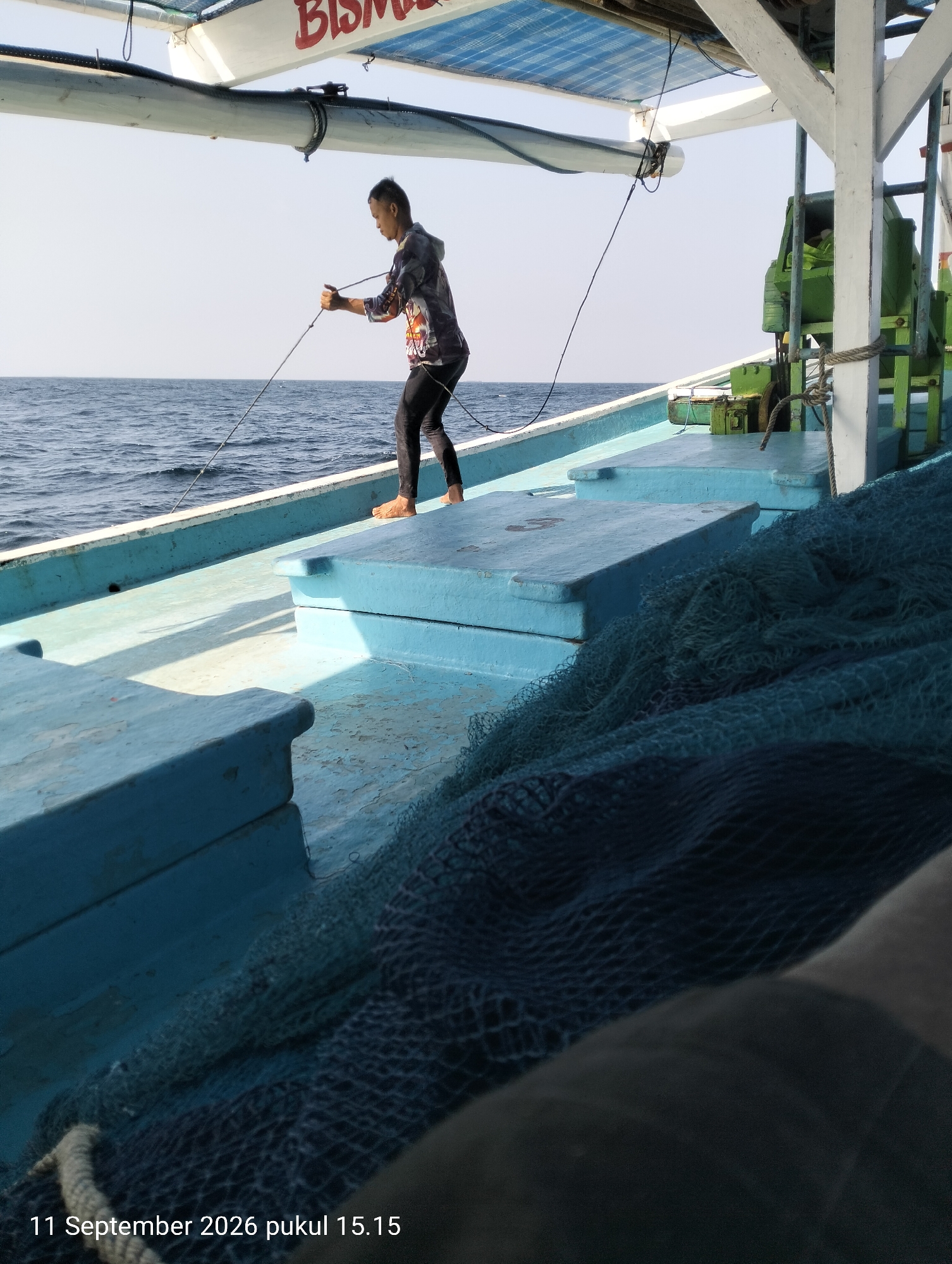 In Bokori, Indonesia, a person stands on a blue boat deck, holding a rope taut. The boat is on the ocean under a clear sky. The person is wearing a patterned hoodie and black pants, with bare feet. A fishing net is piled on the deck of the boat. The boat has a white canopy with red lettering that is partially visible. There is a green engine or equipment on the boat. The timestamp "11 September 2026 pukul 15.15" is overlaid on the bottom of the image.