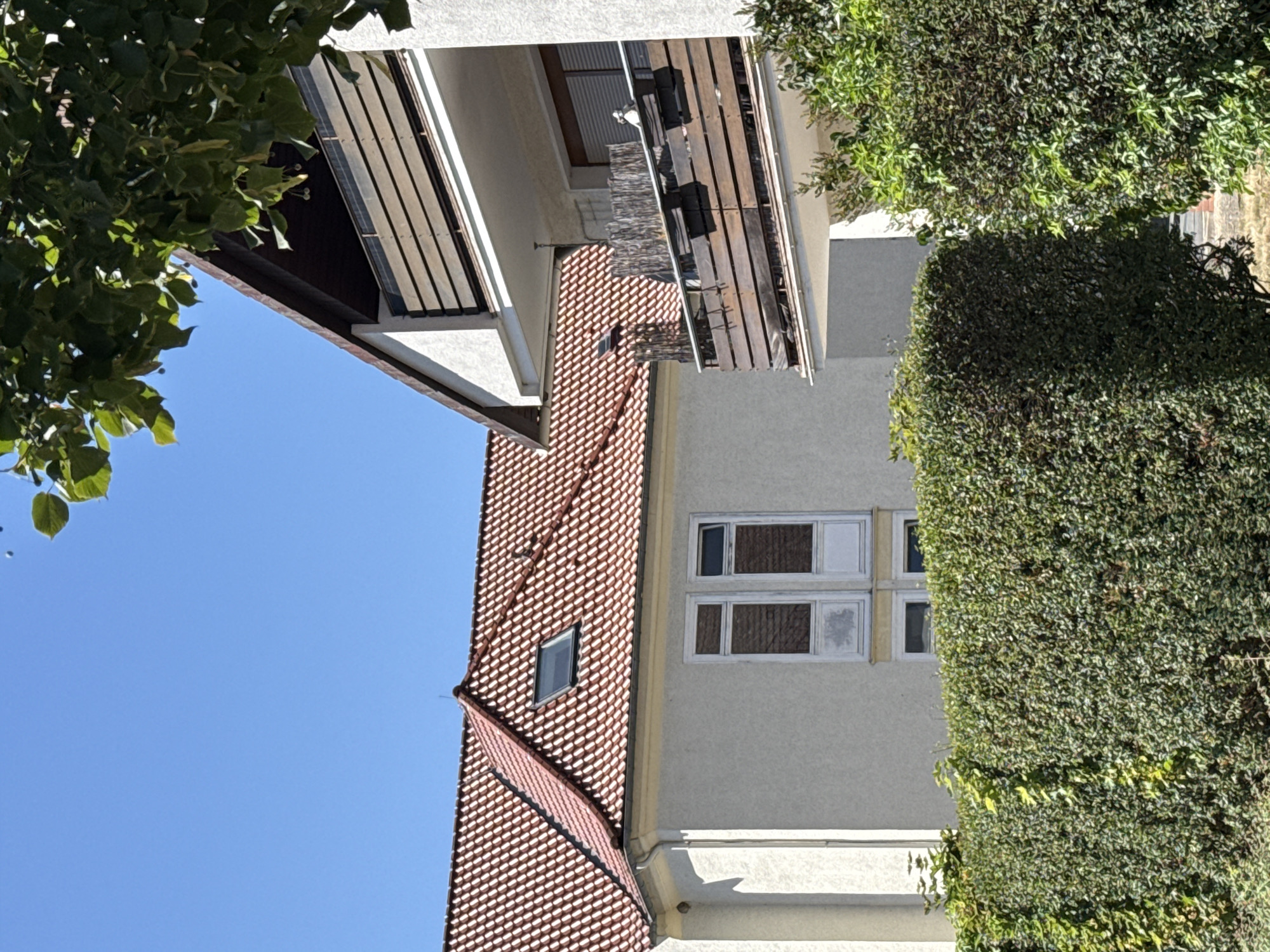 The image shows a residential building with a red tiled roof and a light-colored facade, featuring a skylight. To the right, a modern balcony with wooden railings overlooks a dense green hedge. The sky is clear blue, and a tree with green leaves partially obscures the view of the building's roof. The scene is set in Bad Lauterberg im Harz, Germany.