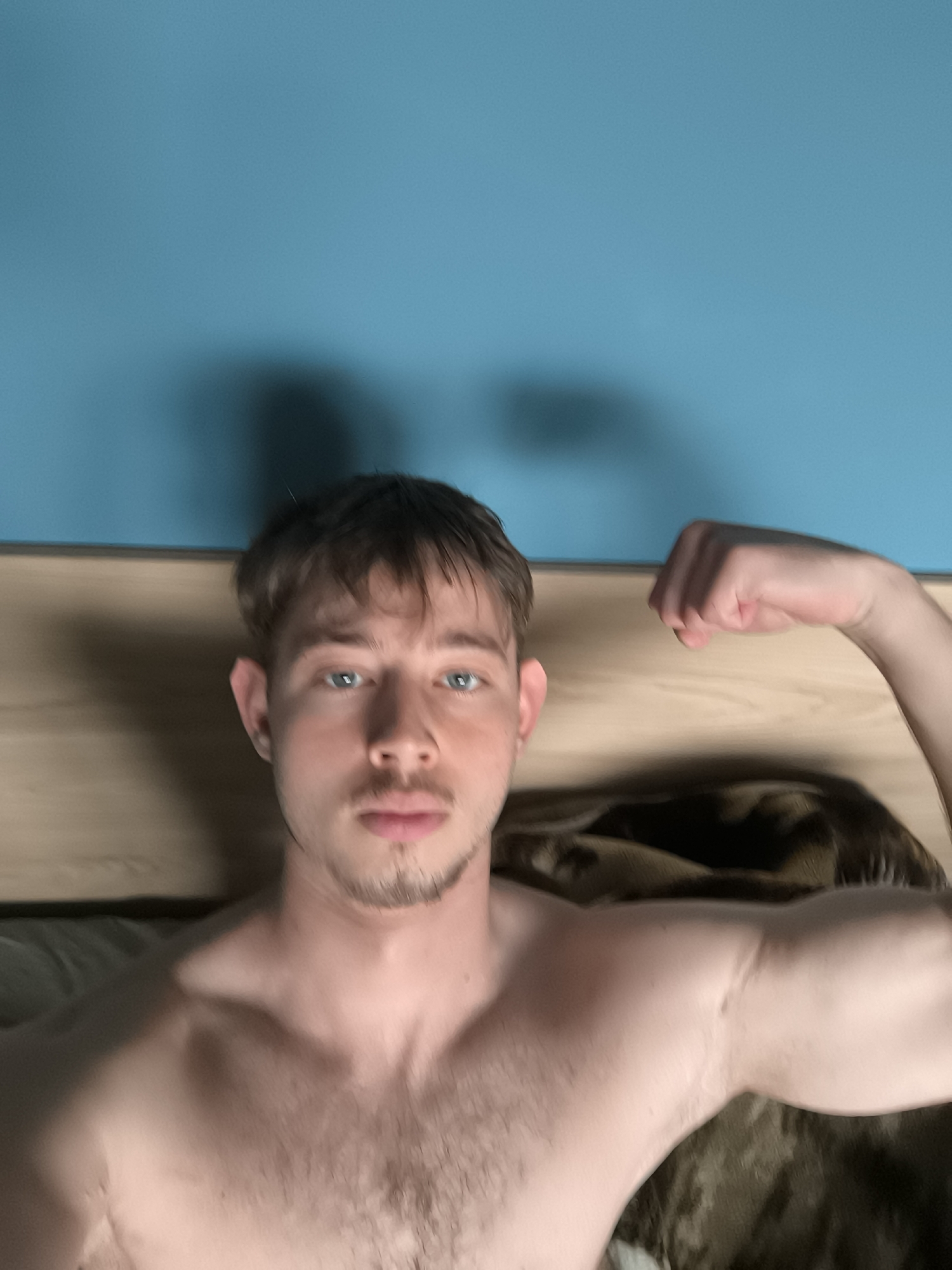 A bare-chested man with light skin and blue eyes is posing indoors. He has brown hair, a short beard, and is flexing his right arm. Behind him is a light-colored headboard and a blue wall. A dark-colored blanket or comforter is visible in the background. The location is likely indoors, but no specific details are available to pinpoint the scene to Sofia, Bulgaria.