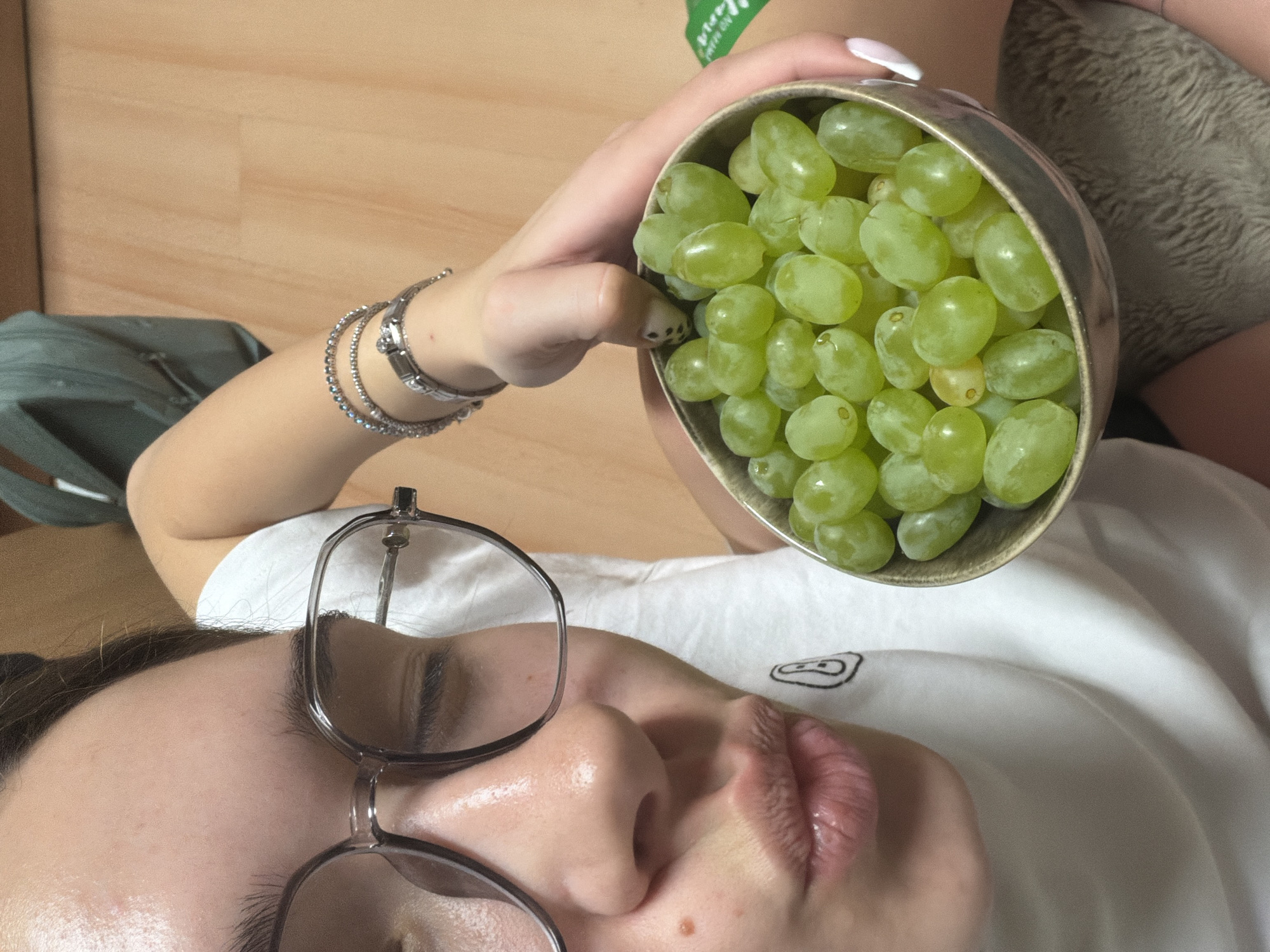 A person lies on a wooden floor wearing a white shirt and glasses. They are holding a bowl of green grapes above their face. A silver bracelet adorns their wrist. There is a green bag in the background. The location is likely indoors. The image does not provide sufficient information to determine if the person is in Sofia, Bulgaria.