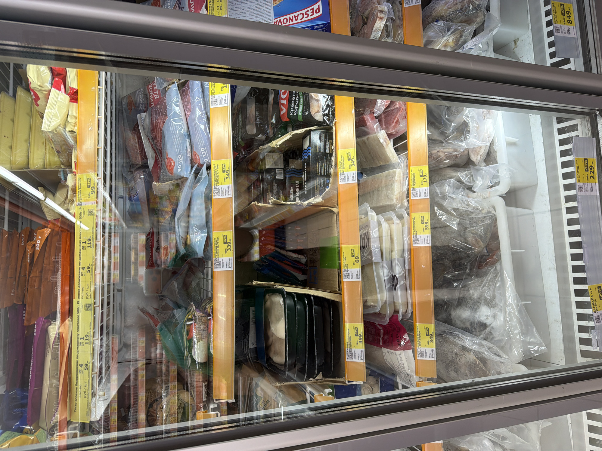The image displays the interior of a commercial freezer unit, likely situated in a grocery store or supermarket, seen through its glass door. The freezer is filled with various packaged frozen food products, predominantly fish, seafood, and dough.
On the left side, numerous yellow packages, possibly of frozen dough or similar items, are stacked. Below them, products labeled "Тесто слоеное бездрожжевое по-домашнему пластовое 500г." (Homemade unleavened puff pastry, 500g) are visible. Further inside, identifiable products include "ФИЛЕ МИНТАЯ" (Pollock Fillet) and "Креветки королевские ОЧИЩЕННЫЕ С" (Peeled King Prawns) from the brand "VICI." Other visible brands are "PESCANOVA," "ПОБРОФЛОТ," and "ВкусАрт," with items like "КАМБАЛА С/м Глазир." (Glazed Flounder) also present. Some products appear stacked in cardboard boxes, indicating a utilitarian display.
Prominently featured throughout the freezer are yellow and white price tags. These labels display product names, original prices, discounted prices, and percentage discounts. Examples include "59 руб./шт" for 4 items of dough and "119 руб./шт" for 1 item, "349" from "499" with "-32%" for "ФИЛЕ МИНТАЯ," and "229" from "299" with "-23%" for "КАМБАЛА С/м." Other price tags show values such as "849" from "1349" with "-37%" and "499" from "799" with "-46%." Most price tags also feature QR codes. The setting is indicative of a retail environment, with artificial indoor lighting. No people or animals are visible.