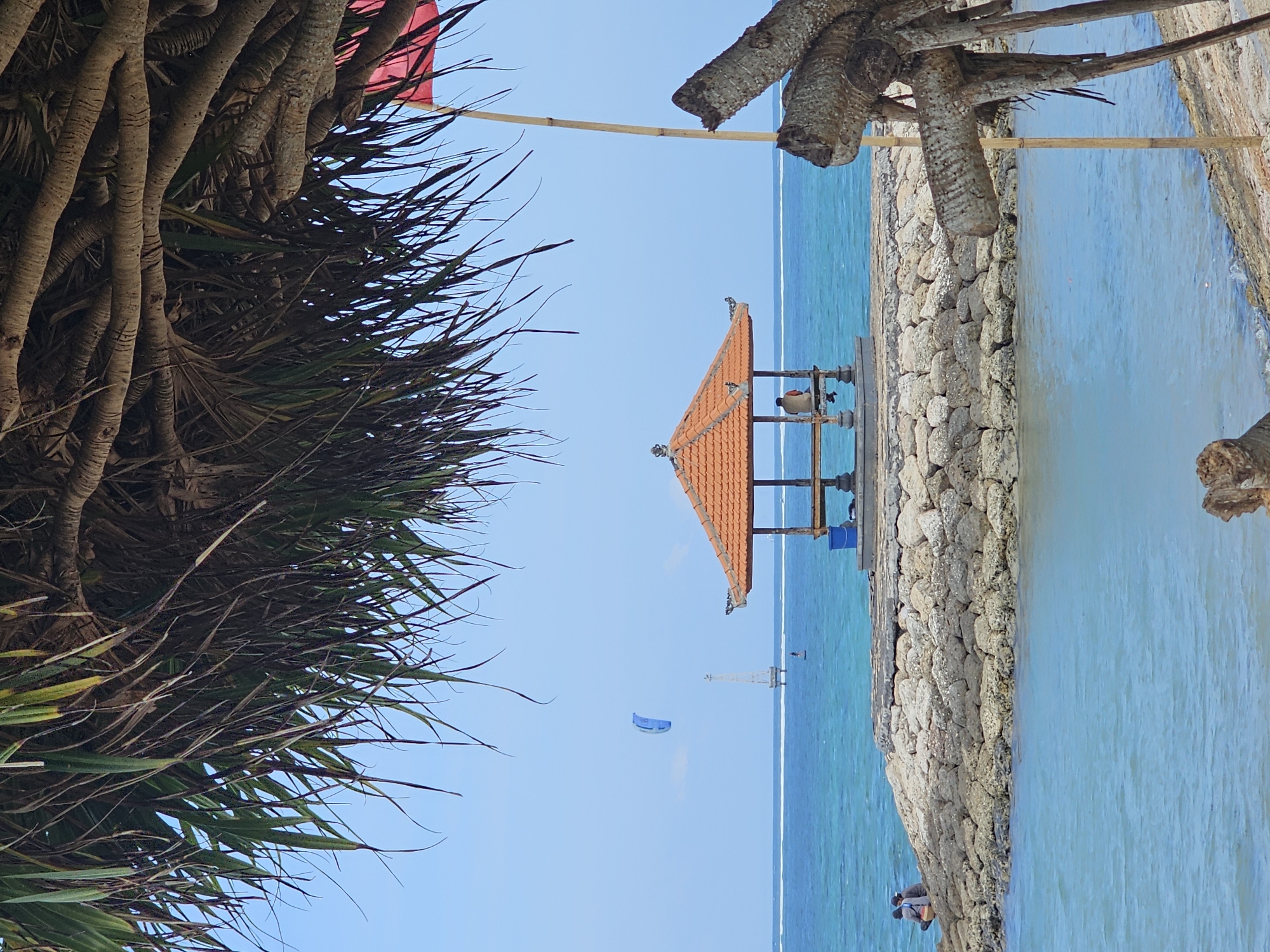 A beach in Denpasar, Indonesia, features a man sitting inside a small, orange-roofed gazebo on a stone jetty. In the distance, a kite surfer and a tall, slender tower are visible on the turquoise ocean under a clear blue sky. The foreground is framed by the drooping fronds of a pandanus tree and a section of driftwood.