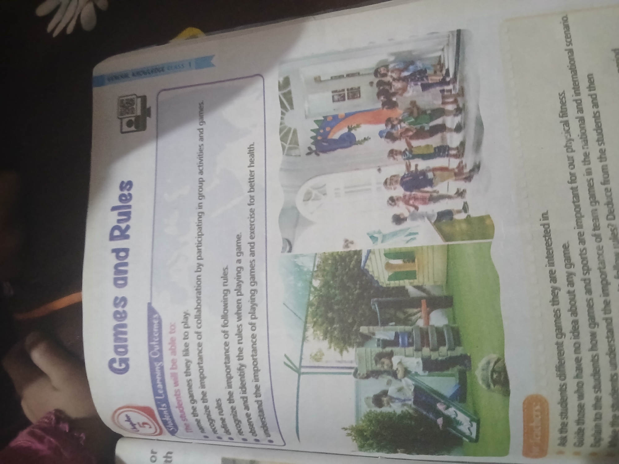 The image shows an open book on a table. The book is titled "Games and Rules" and features a chapter number 5. Below the title, there is a section labeled "Students' Learning Outcomes" which lists several bullet points. The rest of the page is taken up by two photographs. The left photograph shows a playground scene with children and a tortoise. The right photograph shows a group of children standing in a line in front of a colorful mural of a dragon. The book appears to be a children's educational book. There are no discernible elements in the image that suggest the location of Peshawar City Tehsil, Pakistan.